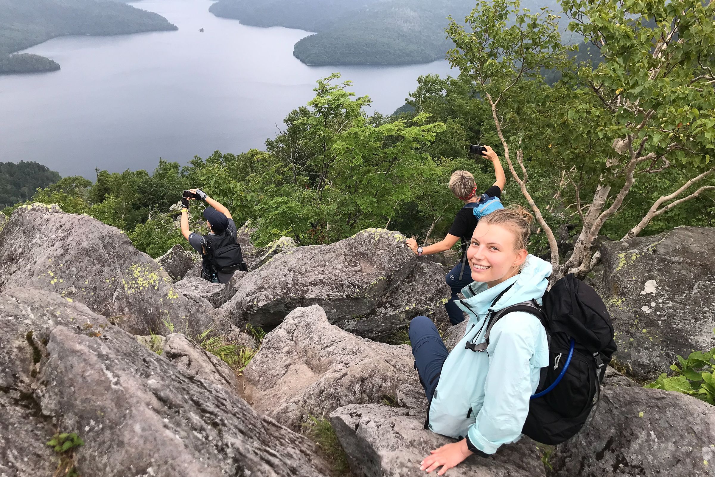 Three hikers sit on boulders atop Mt. Hakuunzan, looking over Lake Shikaribetsu on a cloudy day. One of them in a rain coat smiles at the camera.