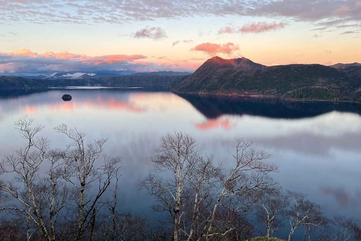 Mt Mashu catches the last light of the day as it is reflected in a glassy Lake Mashu. Leafless birch trees frame the lake in the foreground.