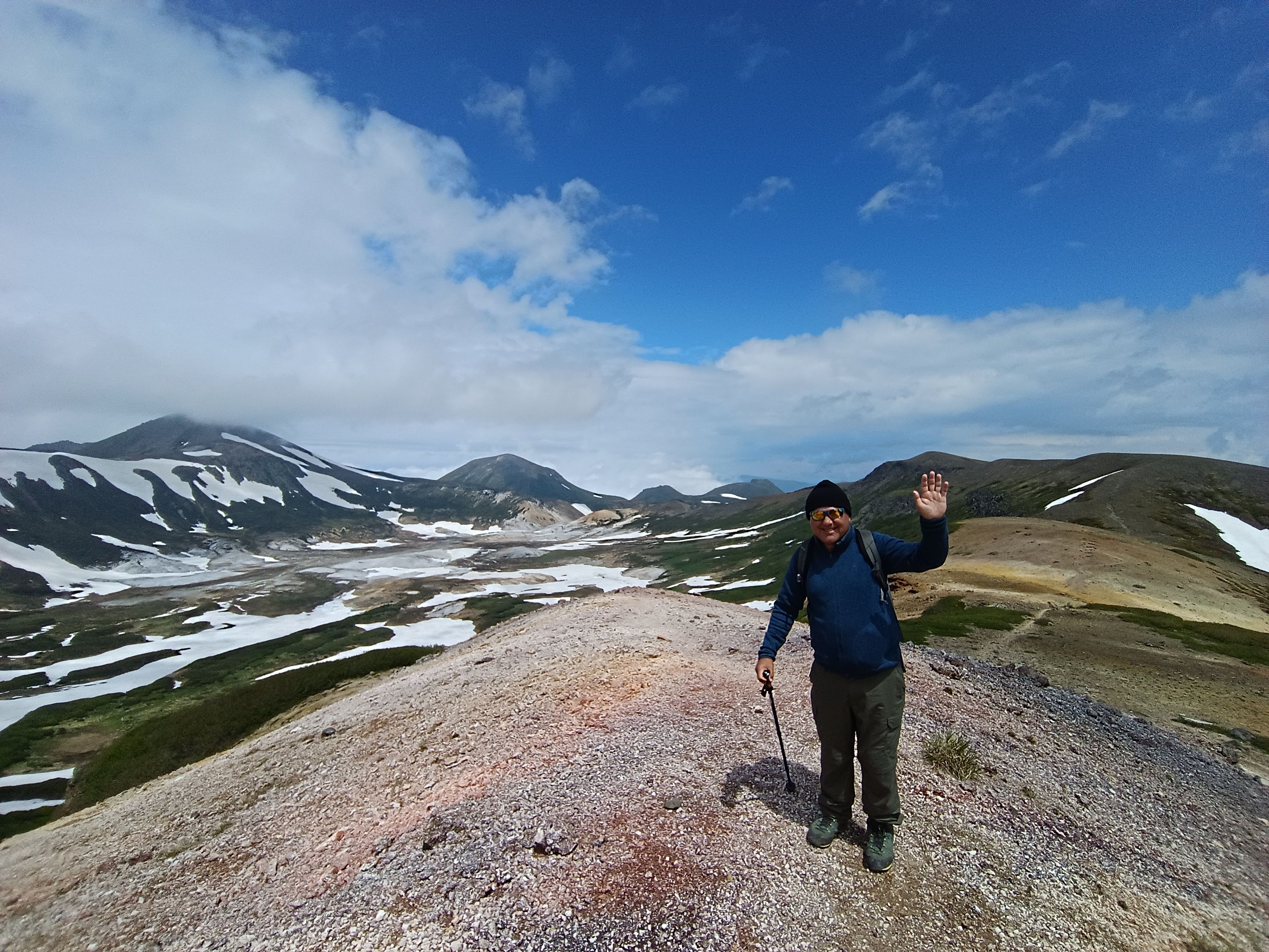 A smiling hiker waves from a rocky ridge, holding a trekking pole. He wears a blue jacket, beanie, and sunglasses. Behind him is a stunning view of the vast Ohachidaira Caldera in Daisetsuzan National Park, featuring rolling mountains, patches of snow, and a bright blue sky with white clouds.