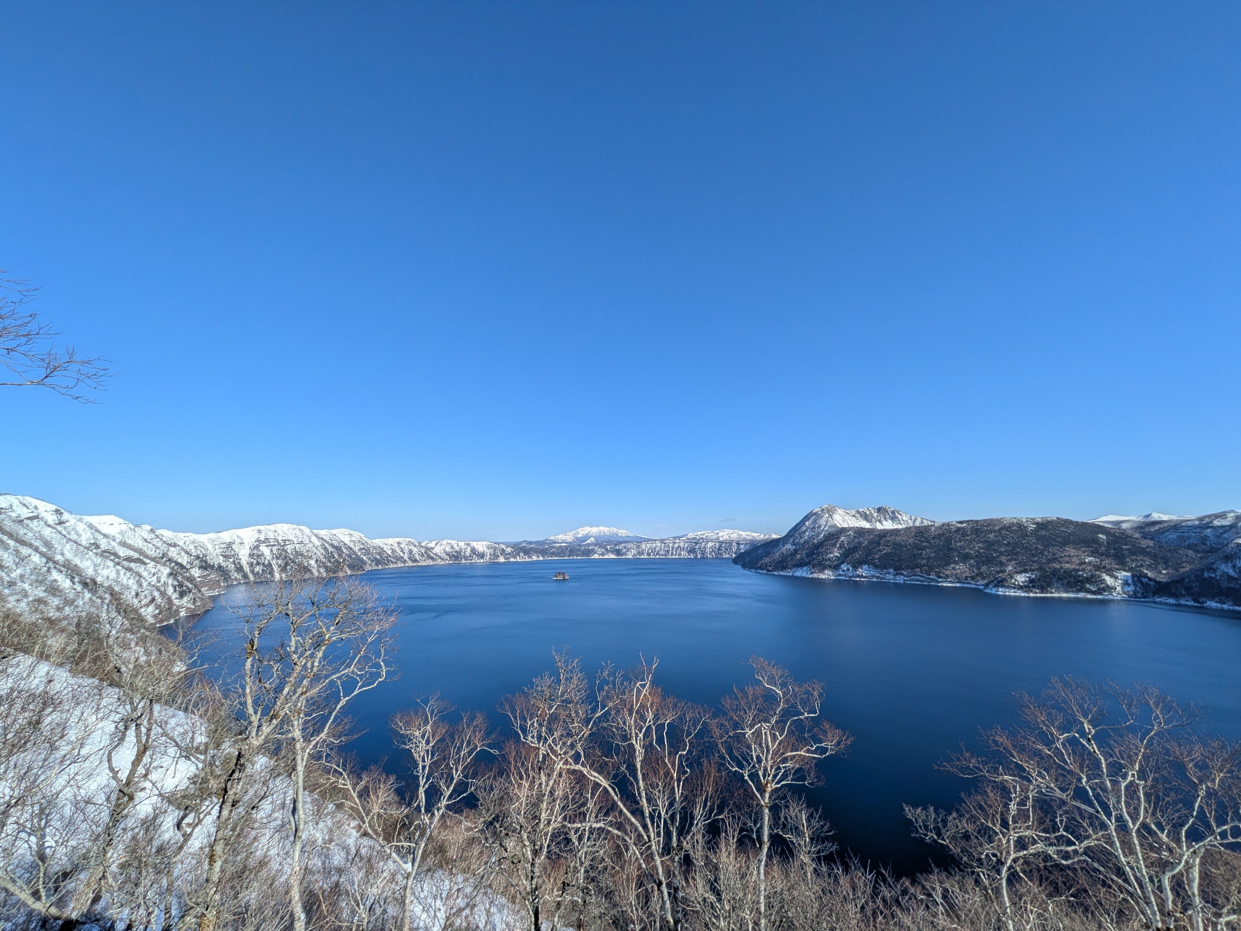 A photograph taken of Lake Mashu, Hokkaido, on a sunny winter day. The sky and surface of the lake are both a pure blue.
