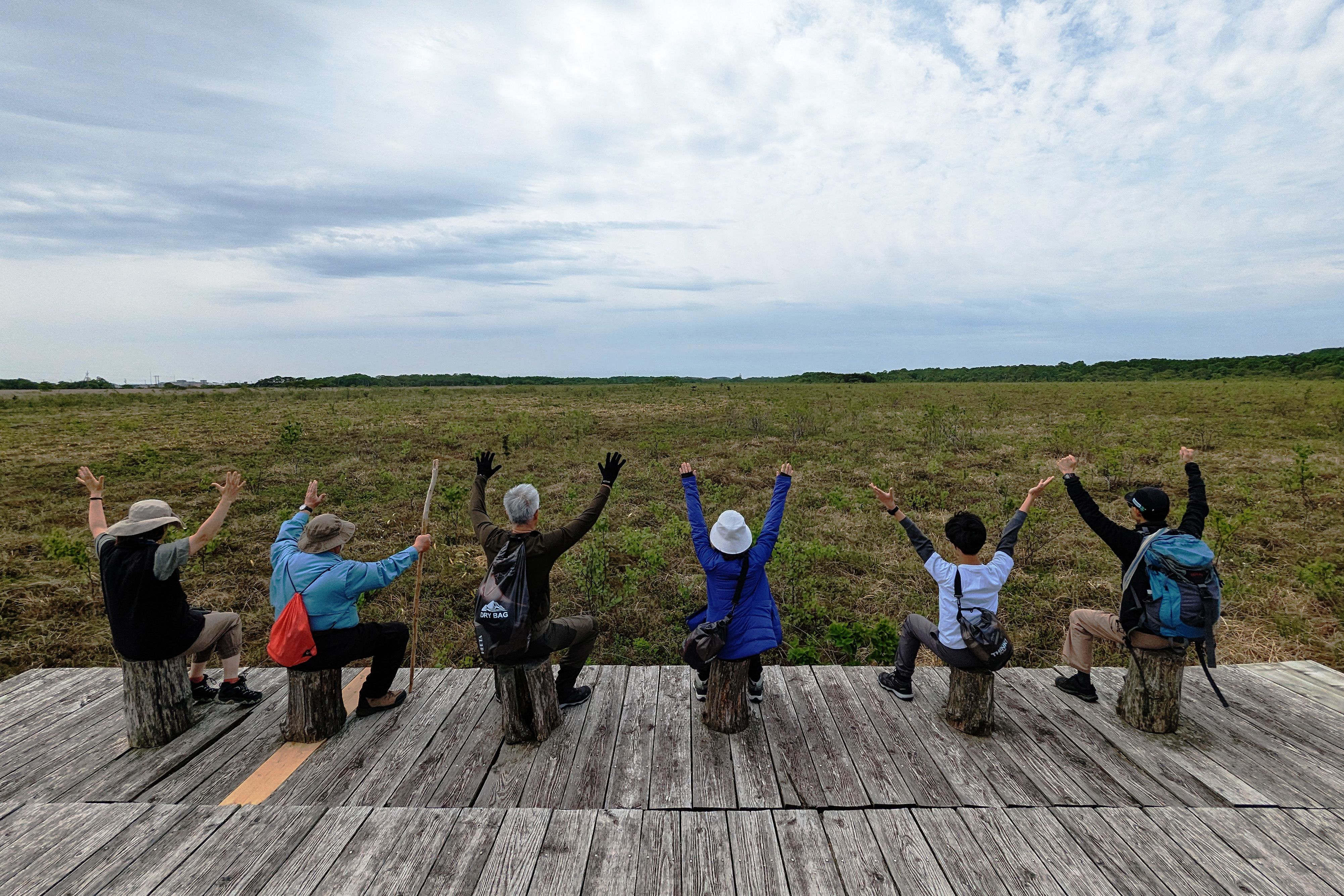 A group of walkers pose for a photo with everyone sat on wooden stools. The group faces away from the camera and towards a wetland