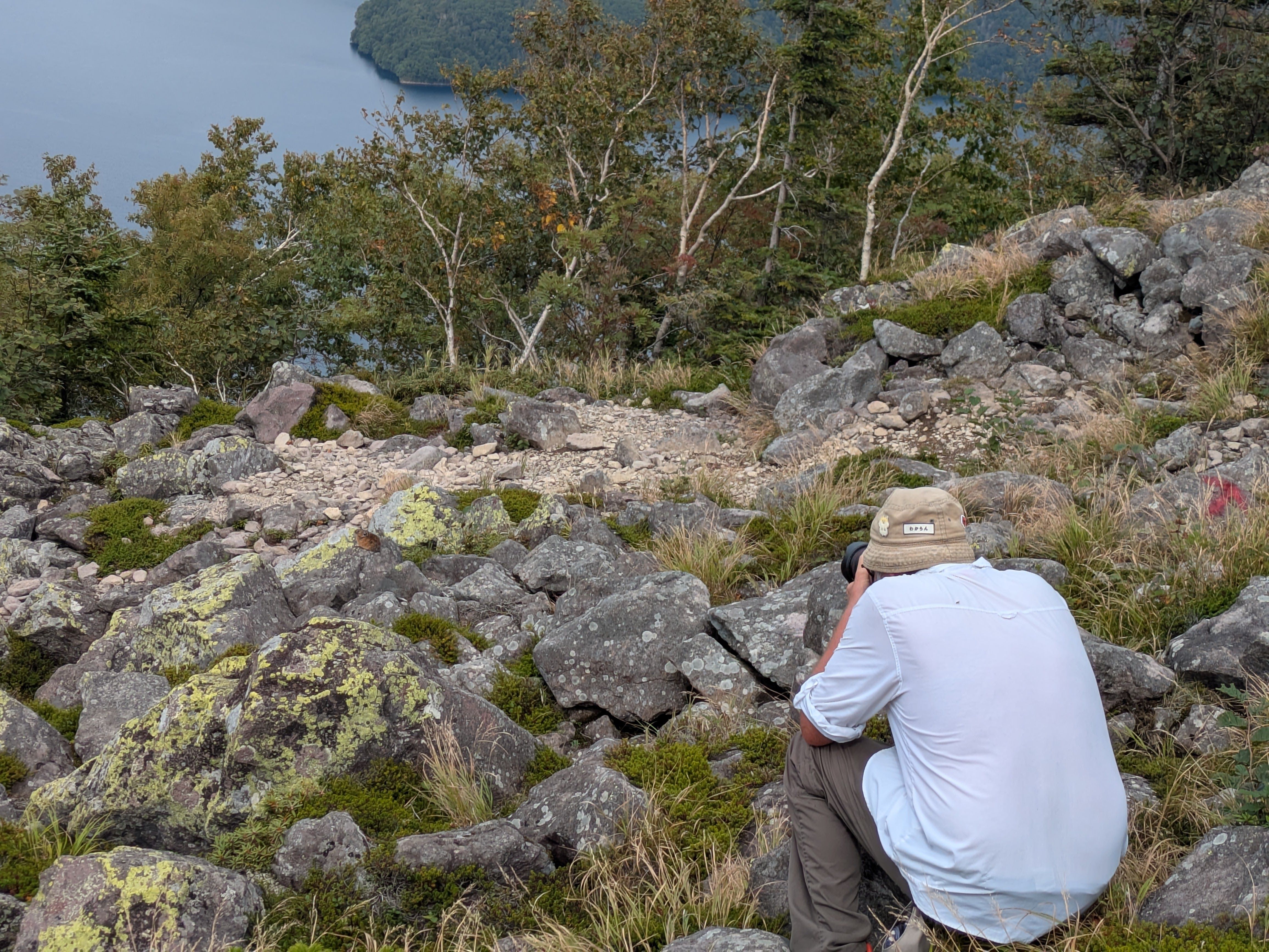 A man crouches in a rock field at the summit of Mt. Hakuun, taking a photograph. The subject is a northern pika, perched atop a rock a few feet away.