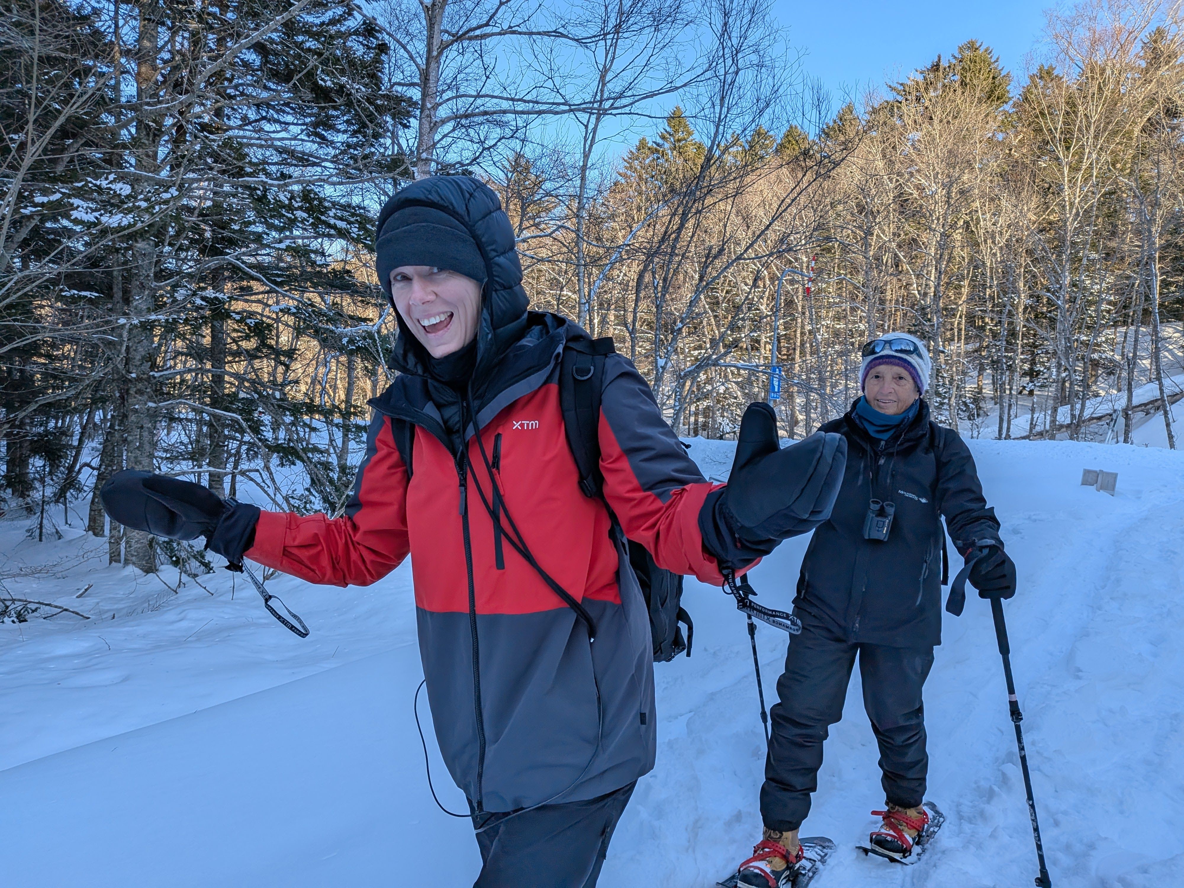 Two women smile at the camera as they snowshoe along a snow-covered road in a forest.