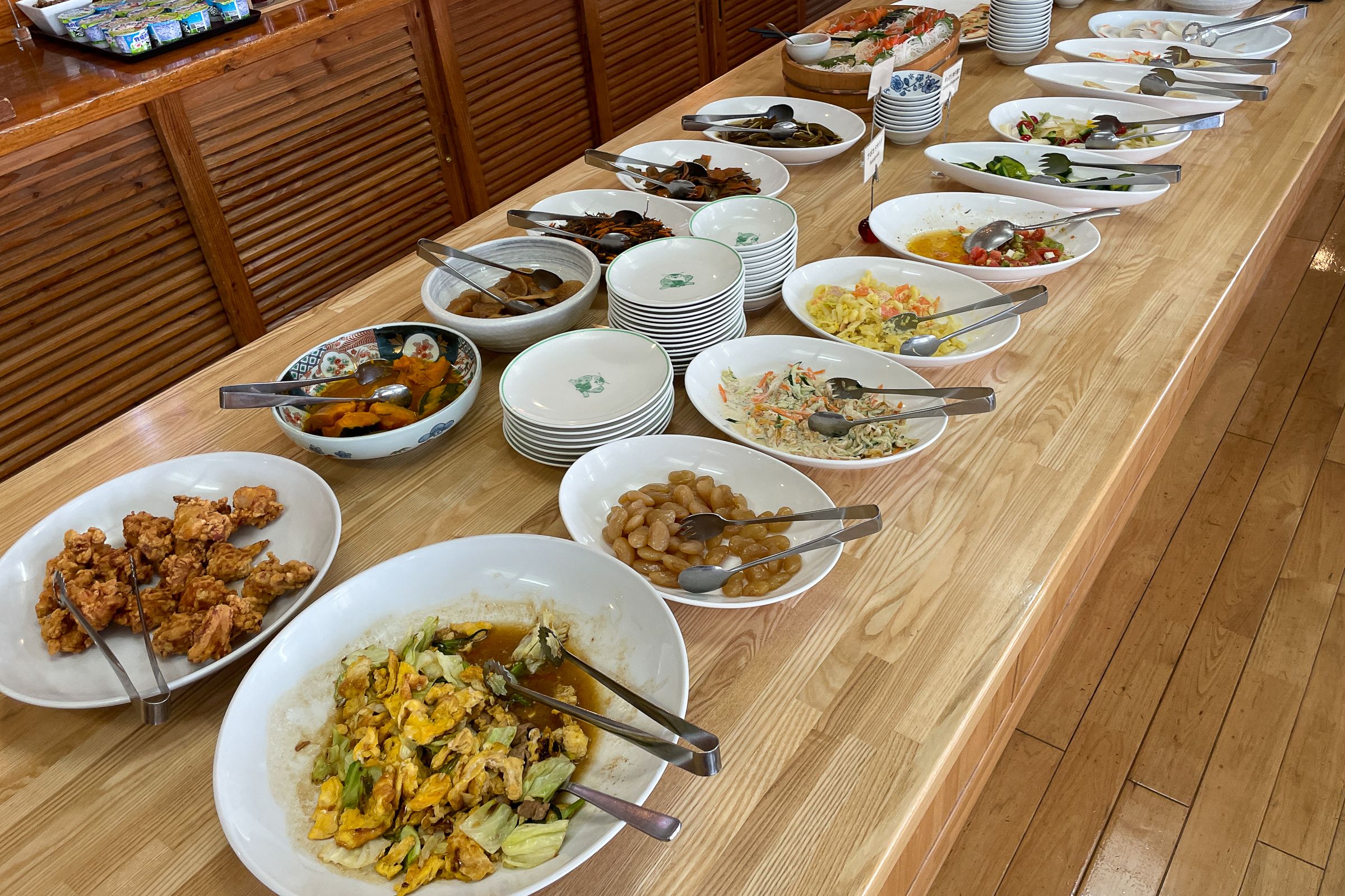 A buffet of colourful and delicious looking food is arranged on a wooden table in two rows of plates. In between the large dishes are small plates and the large dishes have tongs in them.