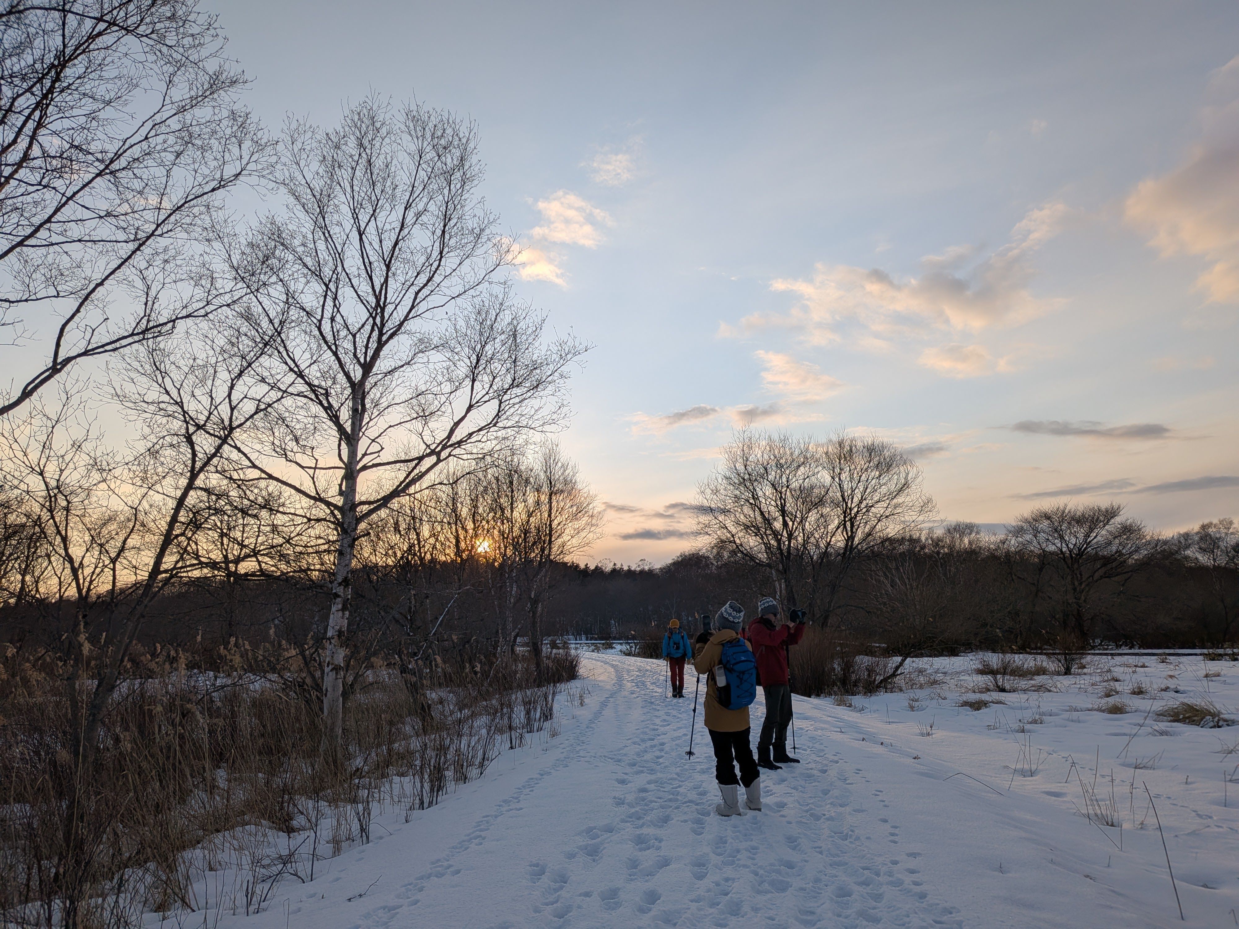 Three people stand on a snow-covered path at sunset, admiring the scenery around them as the light fails. The sky is clear except for a few clouds which reflect the warm evening light.