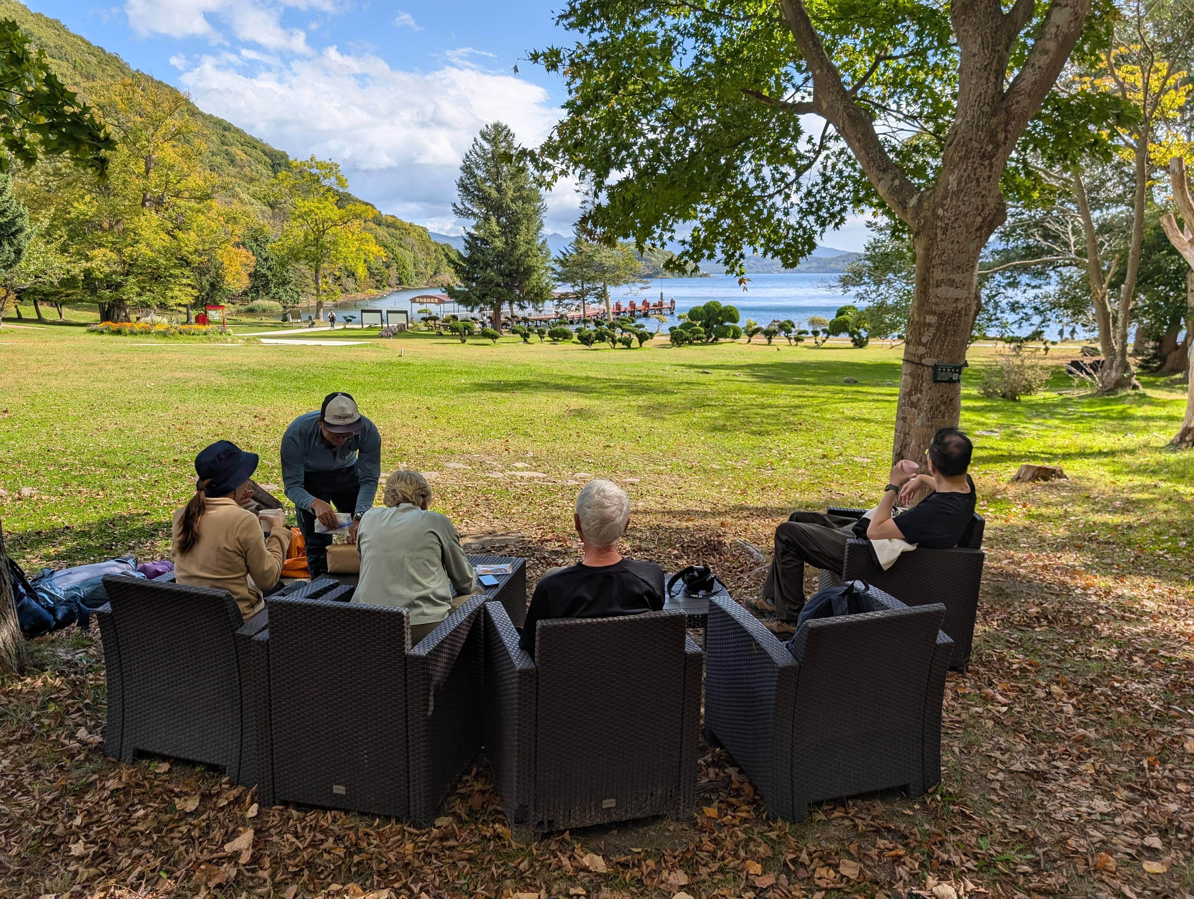 Four hikers are relaxing in dark brown outdoor armchairs on a lawn, scattered with autumn leaves, while waiting for a ferry to Nakajima Island on Lake Toya. A guide pours a drink for a guest. The background features the blue lake and forested hillsides, with boats visible near the shore.