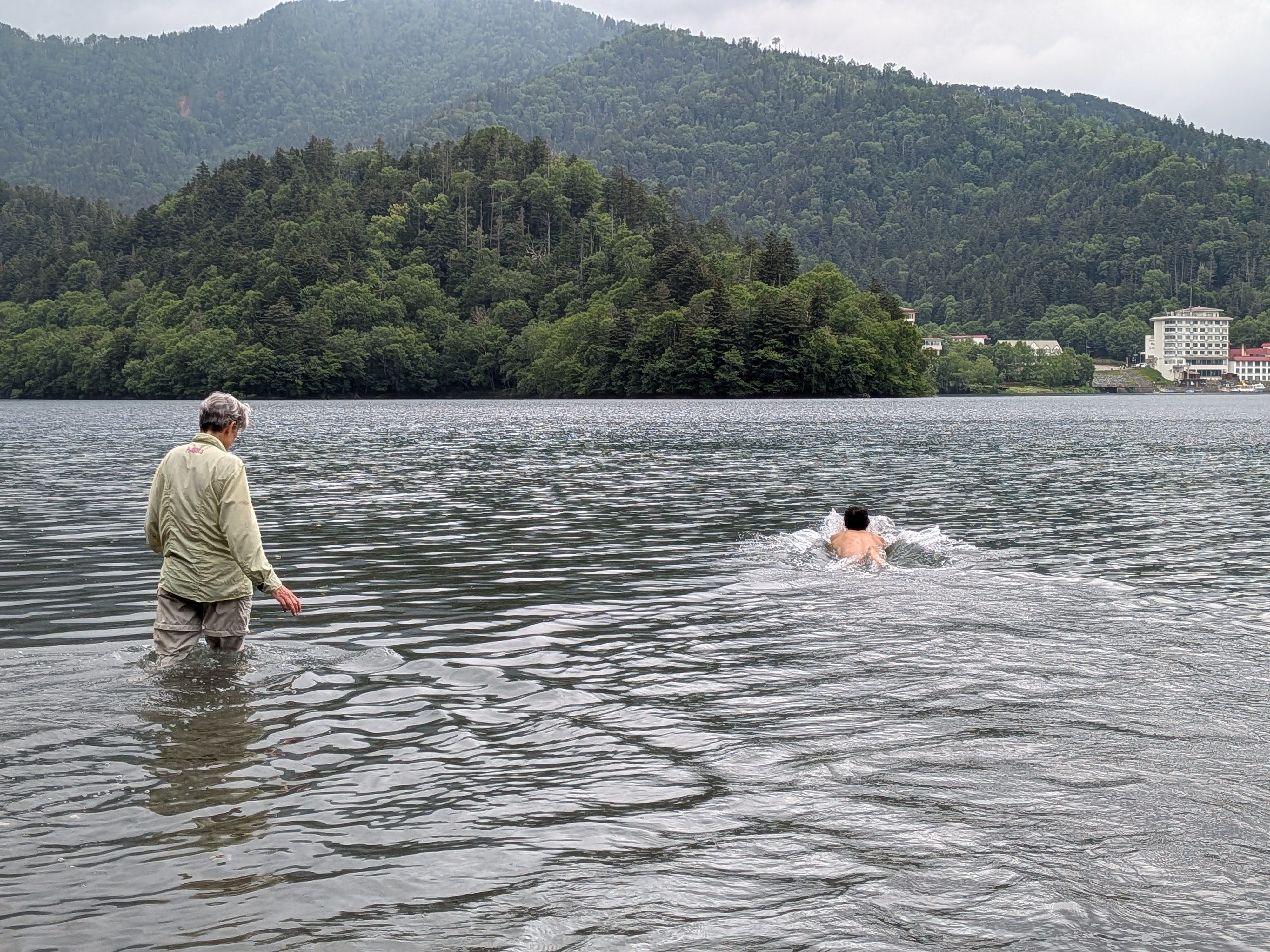 Two people in the water at Lake Shikaribetsu, Hokkaido. One of them is wading in with their clothes on, water to their thighs. The other has removed their shirt and is swimming out into the water.