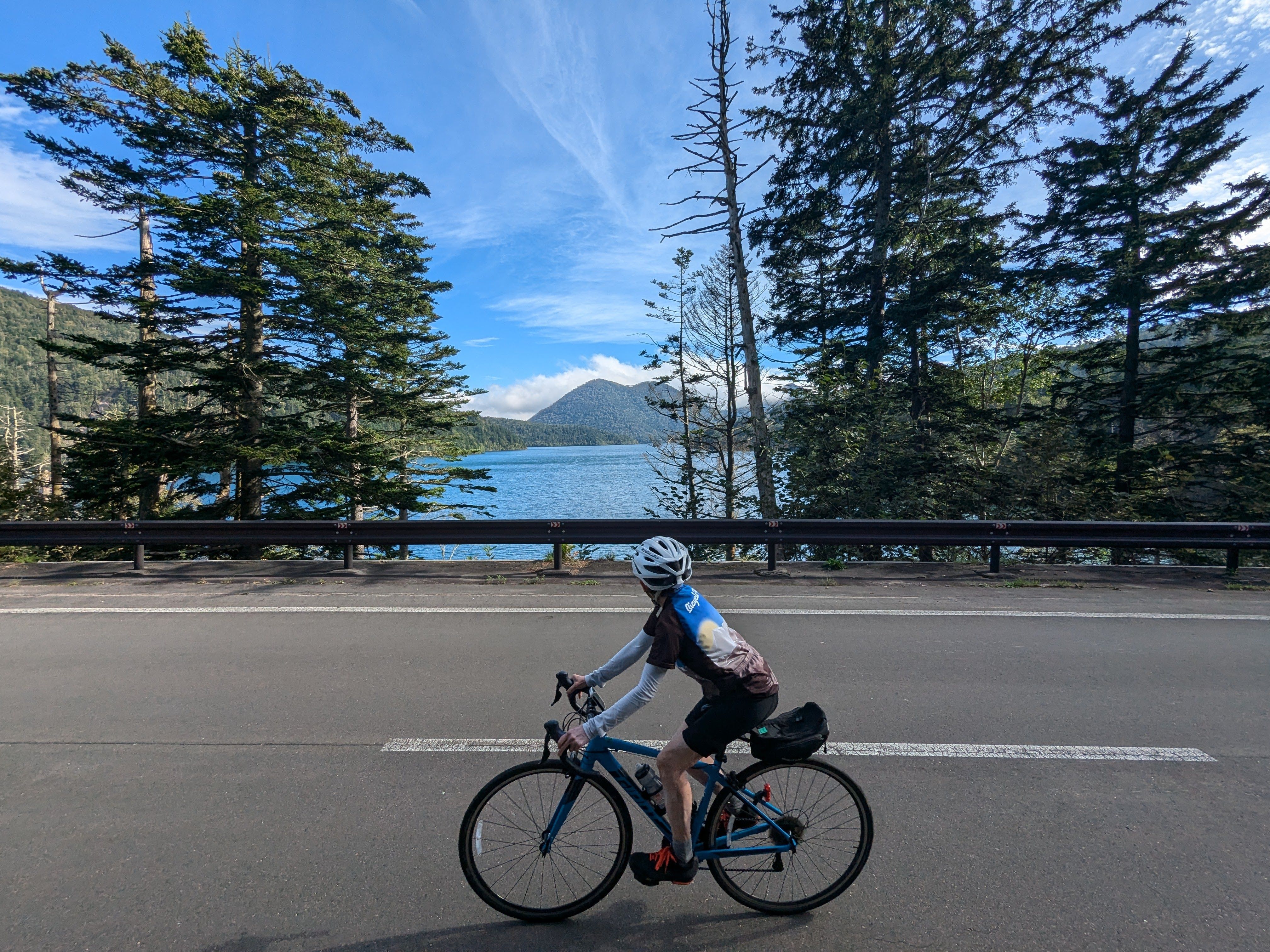 A man on a bike cycles on a road past a lake in the sunshine. The lake and the sky are both bright blue.