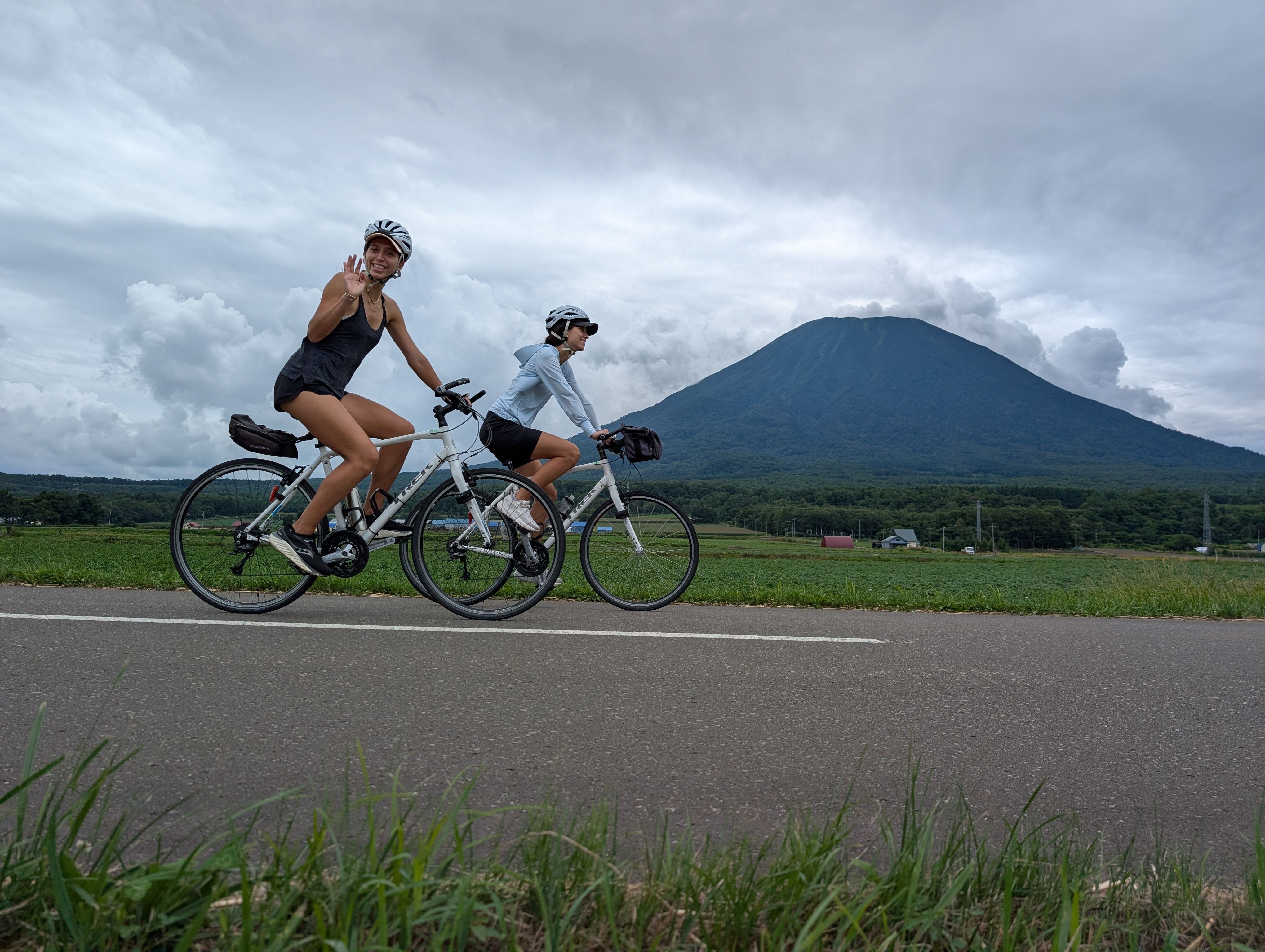 Two cyclists cycle past Mt. Yotei in Hokkaido on a cloudy day. One of the cyclists, a woman, is waving at the camera as she passes.