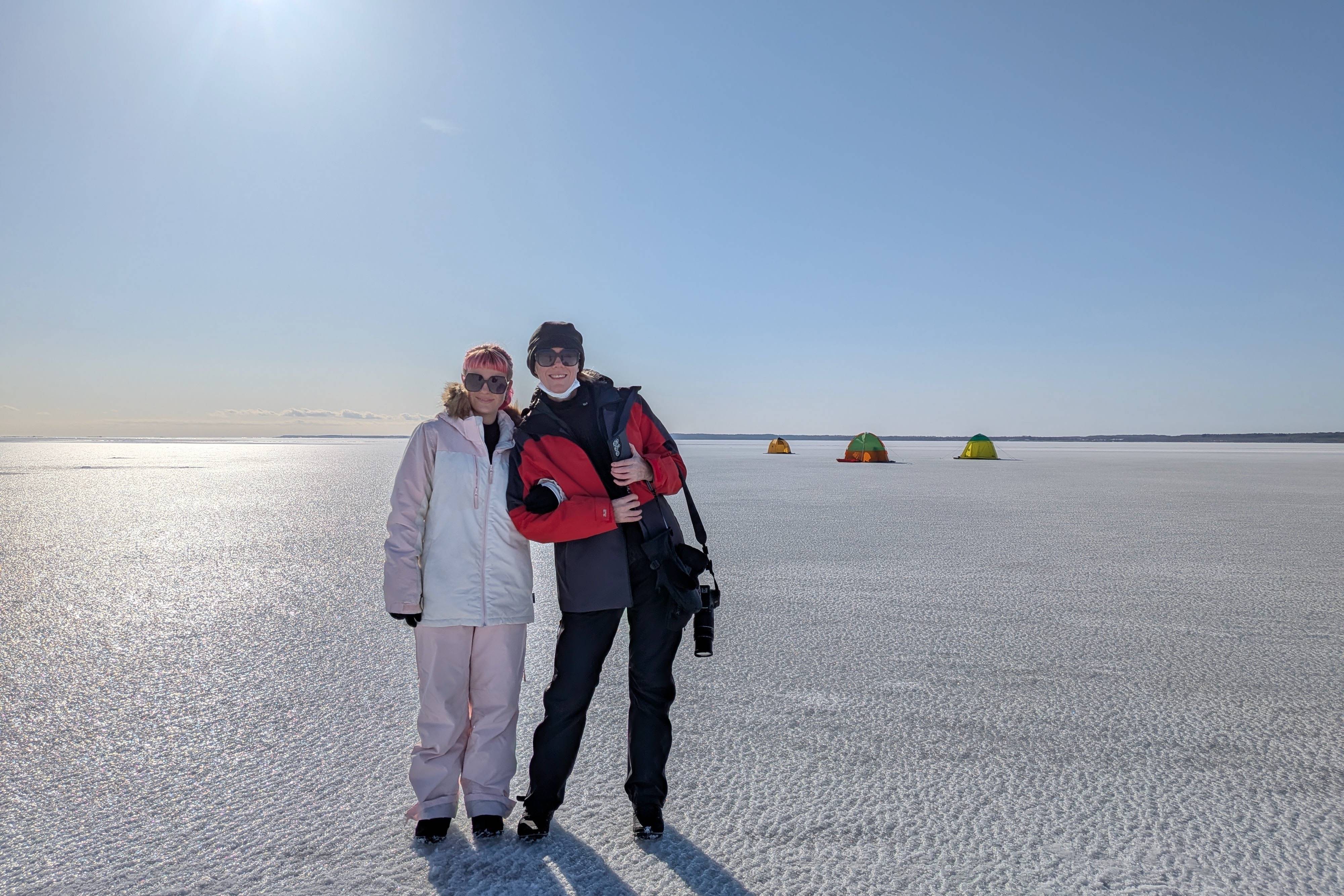 Two smiling guests standing on the expansive frozen surface of Notsuke Bay in Eastern Hokkaido. They are dressed in warm winter gear under a bright blue sky, with colourful ice fishing tents visible in the background.