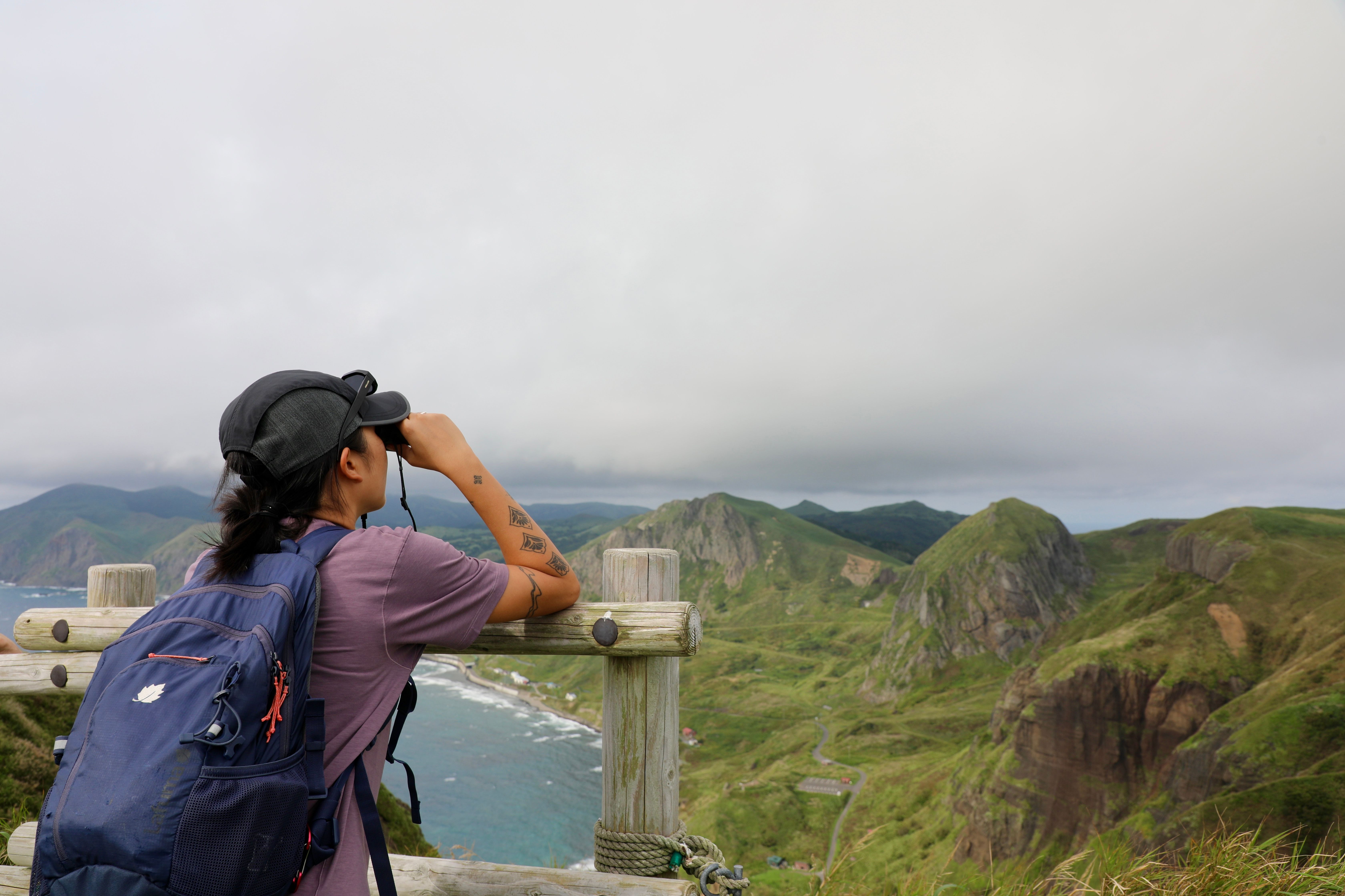 A photograph taken of a woman from behind. She is wearing hiking gear and is leaning on a wooden fence, using binoculars to look out at the view over Rebun Island, Hokkaido. Green mountains and an ocean cape are visible in the distance.