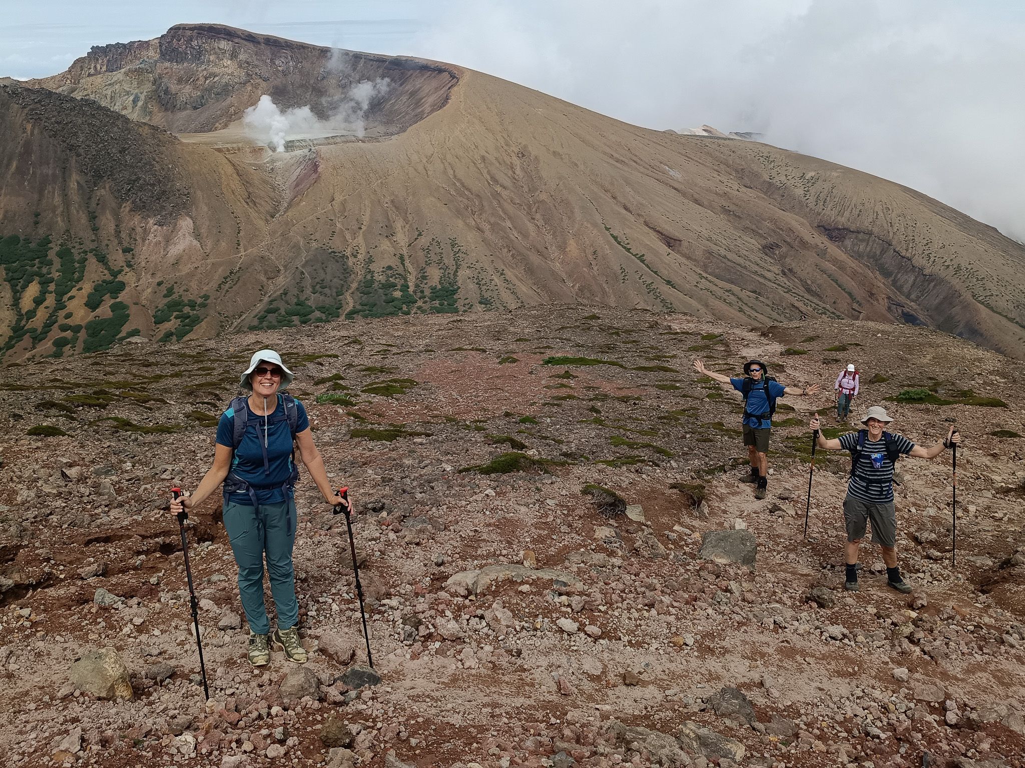 A group of hikers smile at the camera on a mountain trail. A mountain crater steams in the background.