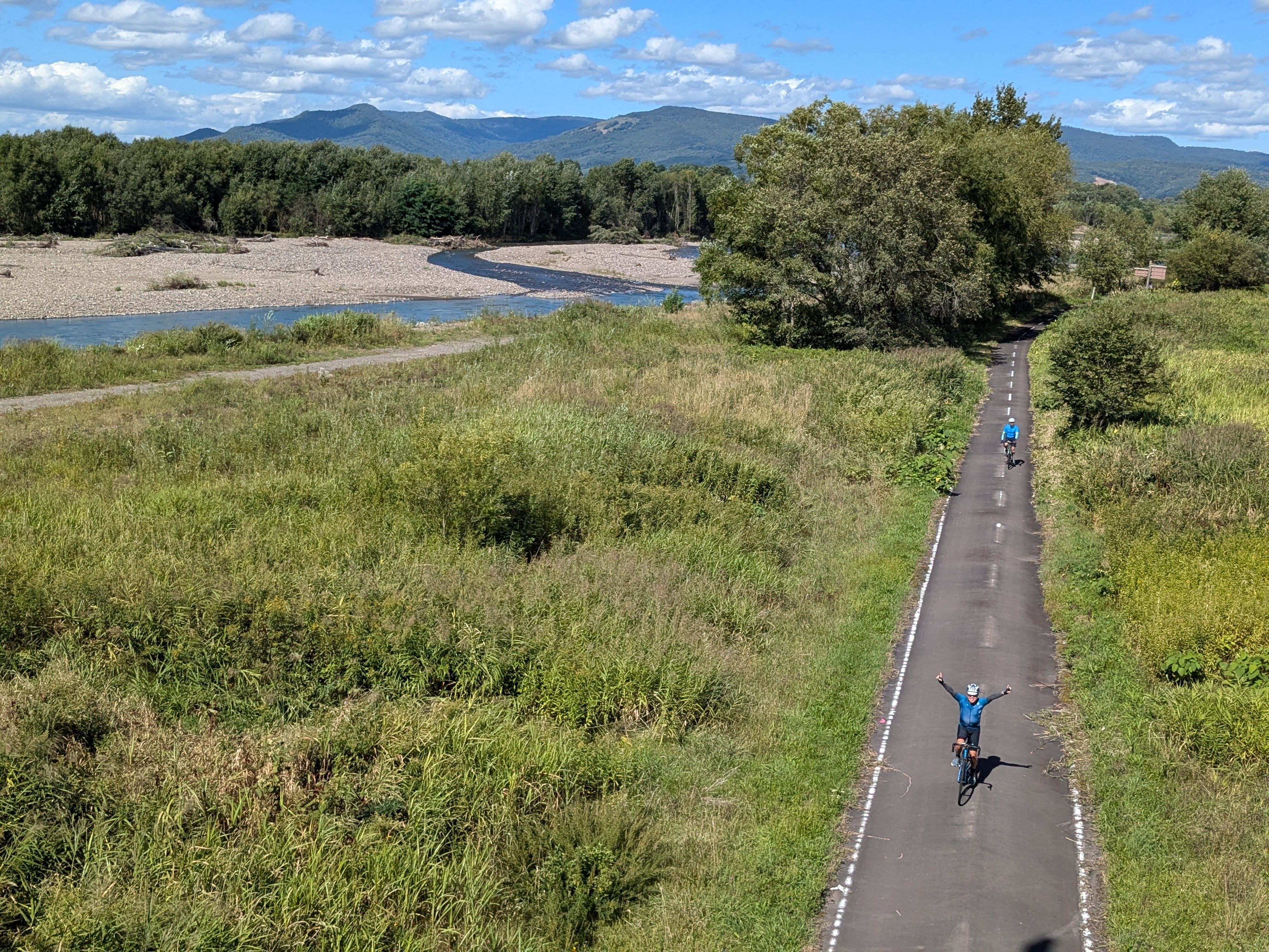 A group of cyclists ride alongside the Ishikari River in Hokkaido. One of the cyclists raises their arms in victory.