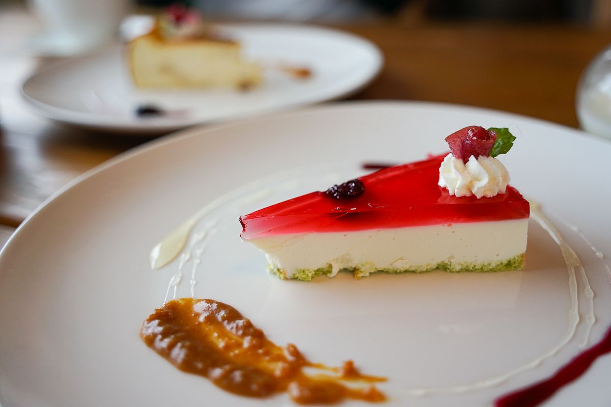 A photograph of a no-bake cheesecake on a plate with various sauces swirled around it. Atop the layer of creamy cheese is a thin layer of berry jelly. It has a green biscuit base, possibly pistachio or green tea.