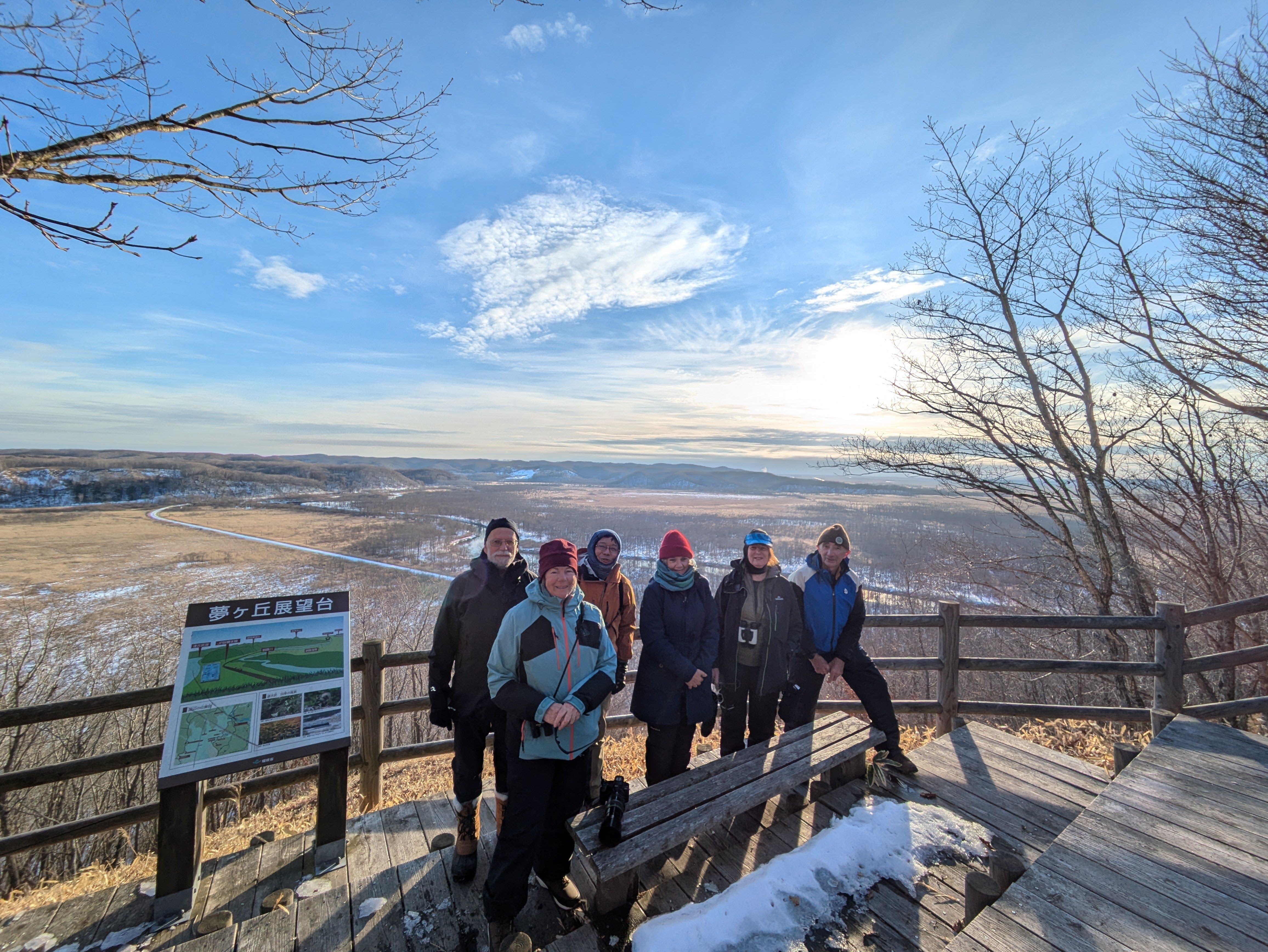 A group of people smile at a camera on a sunny winter day. They are stood on a viewing deck overlooking Kushiro Shitsugen National Park and its vast marshes in Hokkaido.