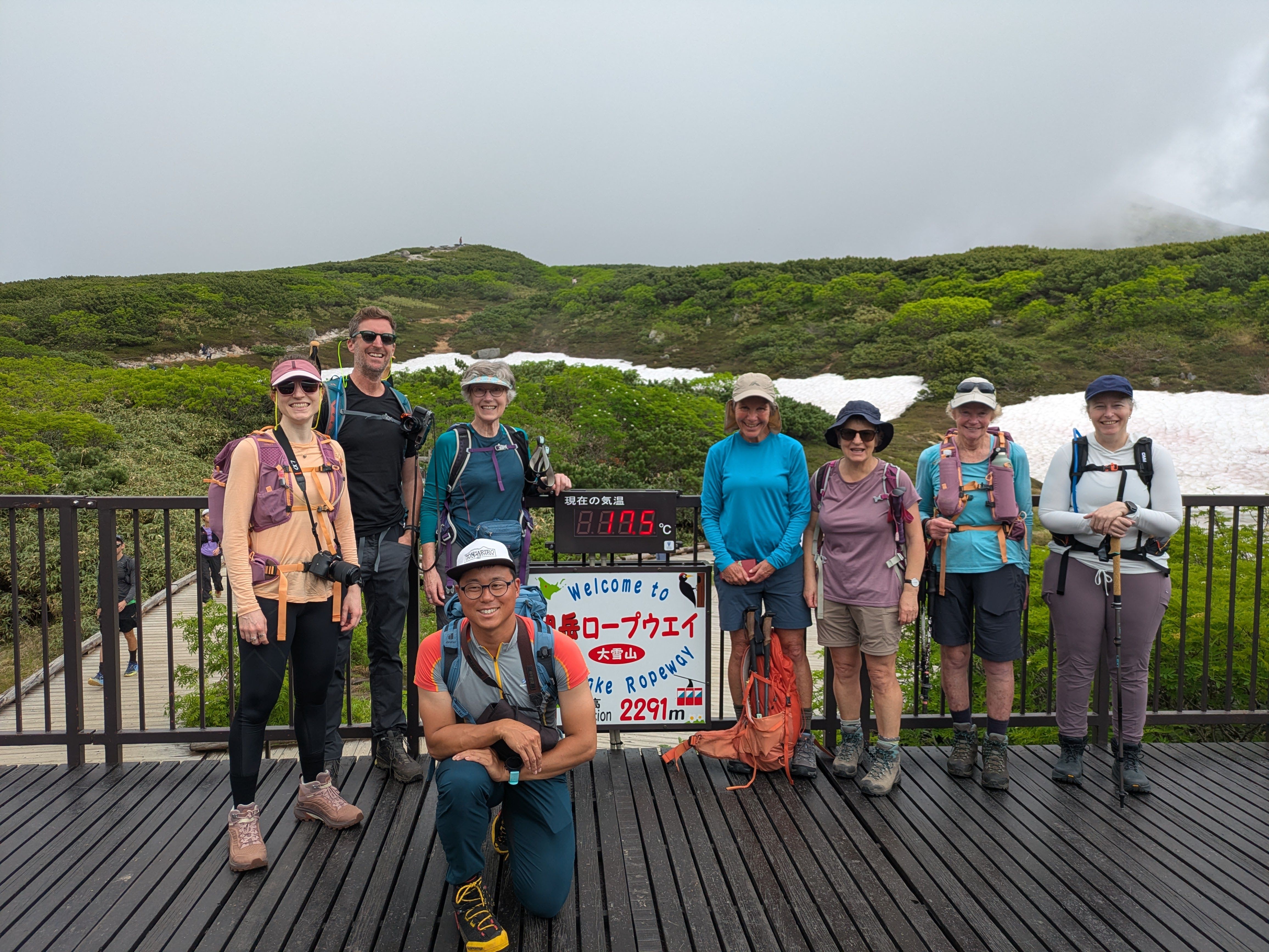 A group of a hikers smile next to a sign at the top of Asahidake Ropeway. It is cloudy, so the mountain behind them is not visible. The sign next to them indicates the group is at 2291m altitude.