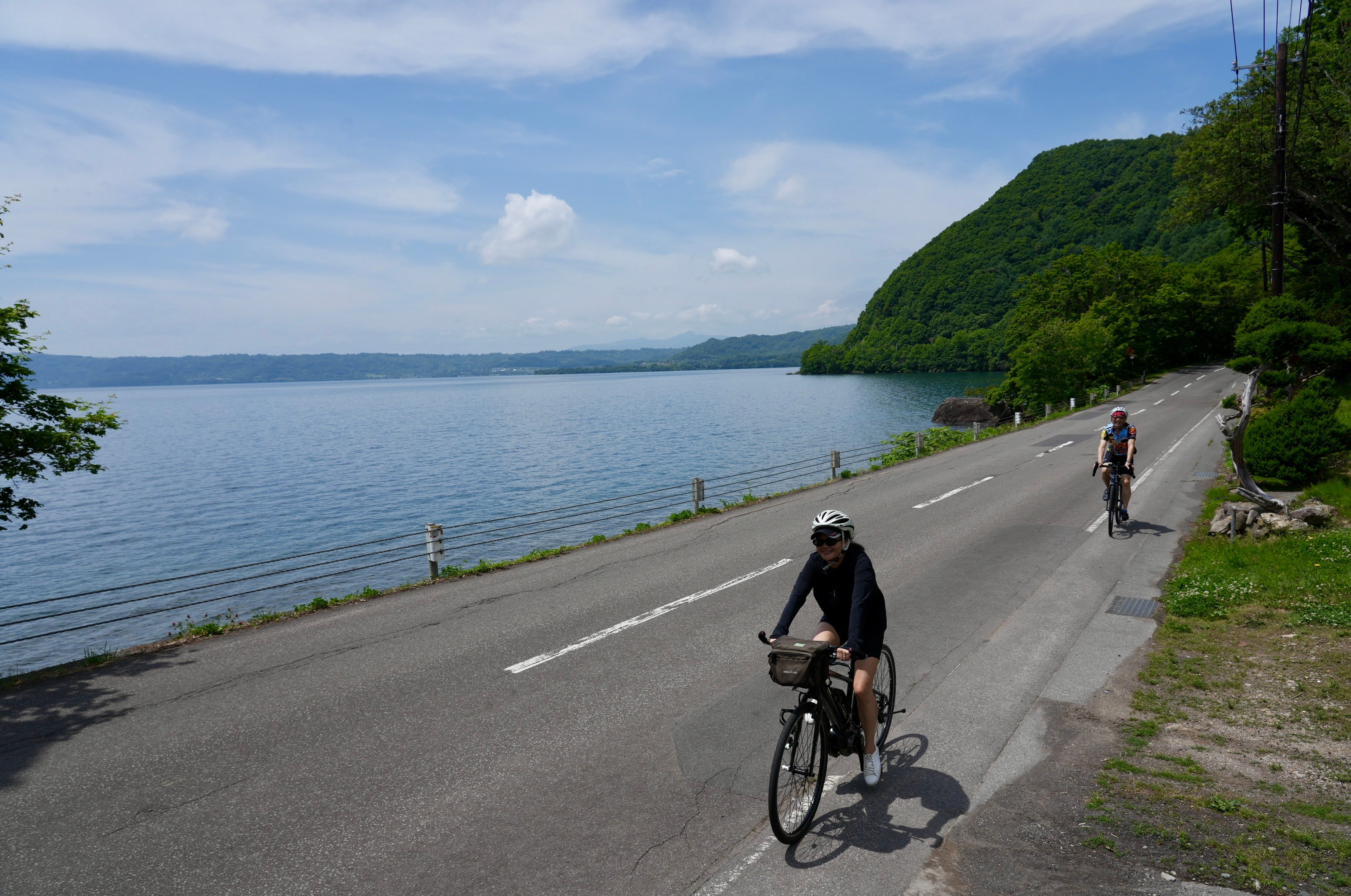Two cyclists ride along a road on the shoreline of Lake Toya, Hokkaido. It is a sunny day and both the sky and the waters of the lake are blue.