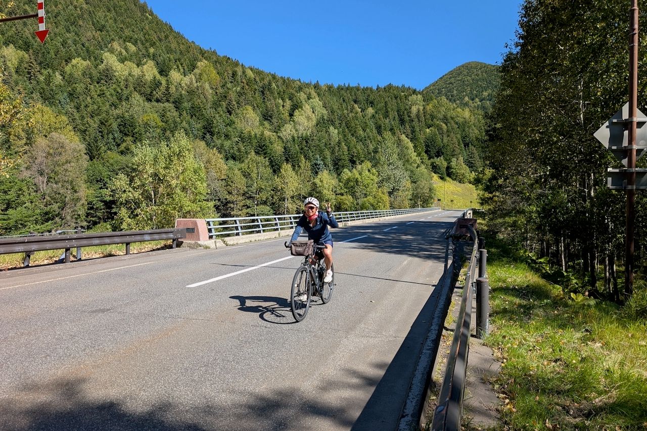 A happy cyclist flashes a peace sign while riding her bike across a bridge on a sunny day. She is descending from Mikuni Pass towards Lake Taisetsu on a paved road. Lush green forested mountains rise in the background under a clear blue sky.