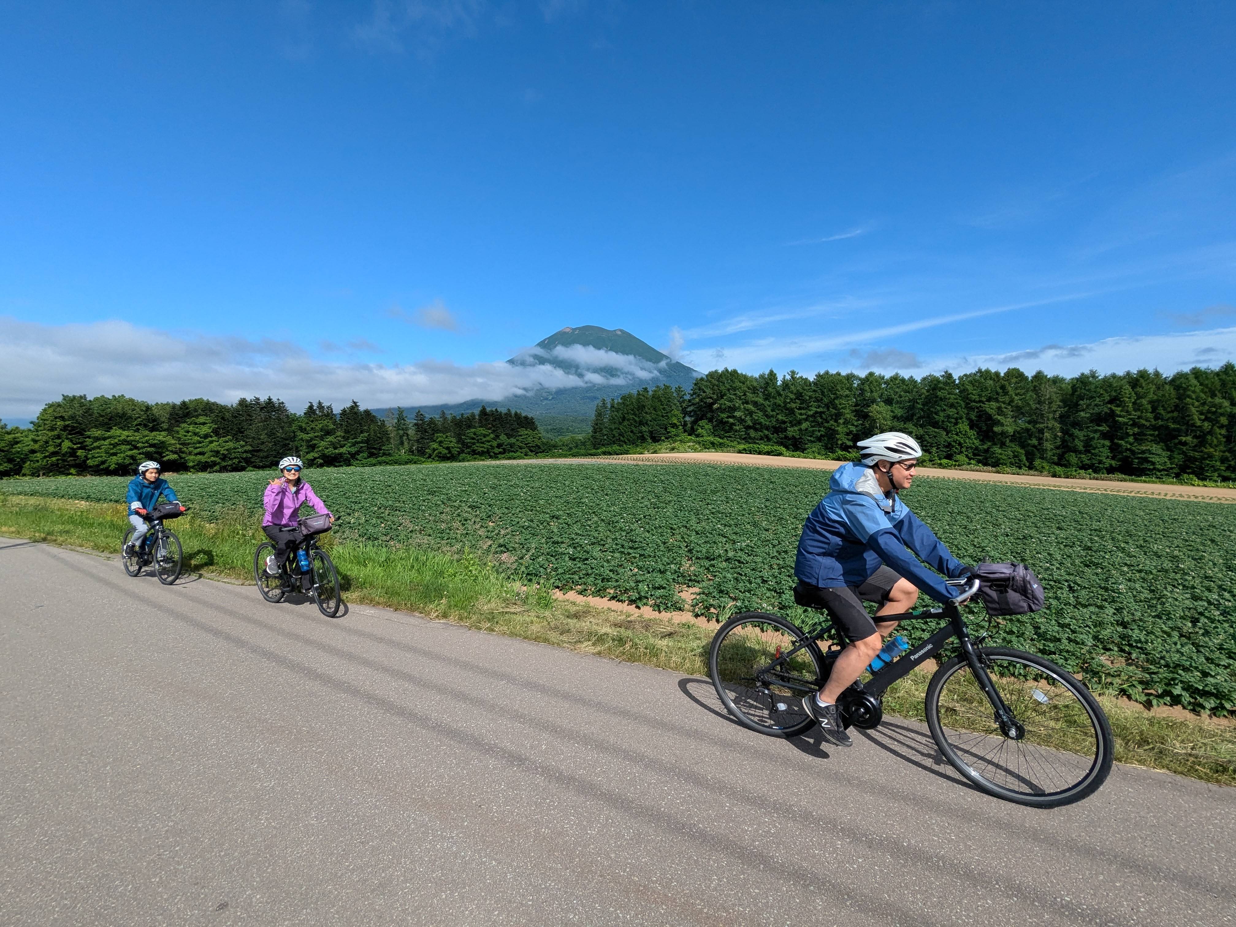 Three cyclists ride in a line past Mt. Yotei in Hokkaido. It is a beautifully sunny day. Mt. Yotei is partially obscured by clouds, giving it a mysterious look.