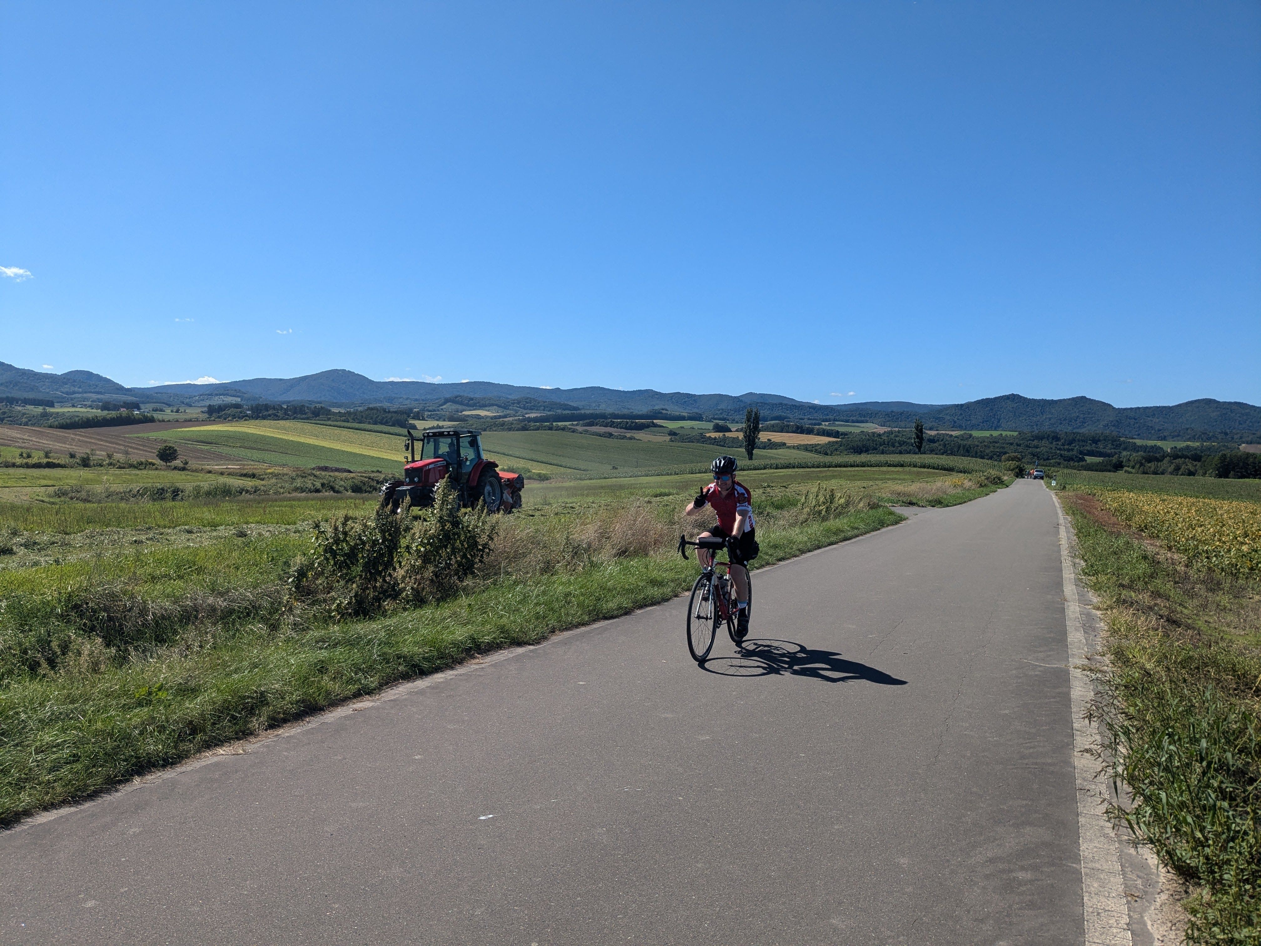 A cyclist rides along a back road in Biei, Hokkaido, alongside two agricultural fields. There's a tractor in the field next to him. It is a beautiful sunny day.