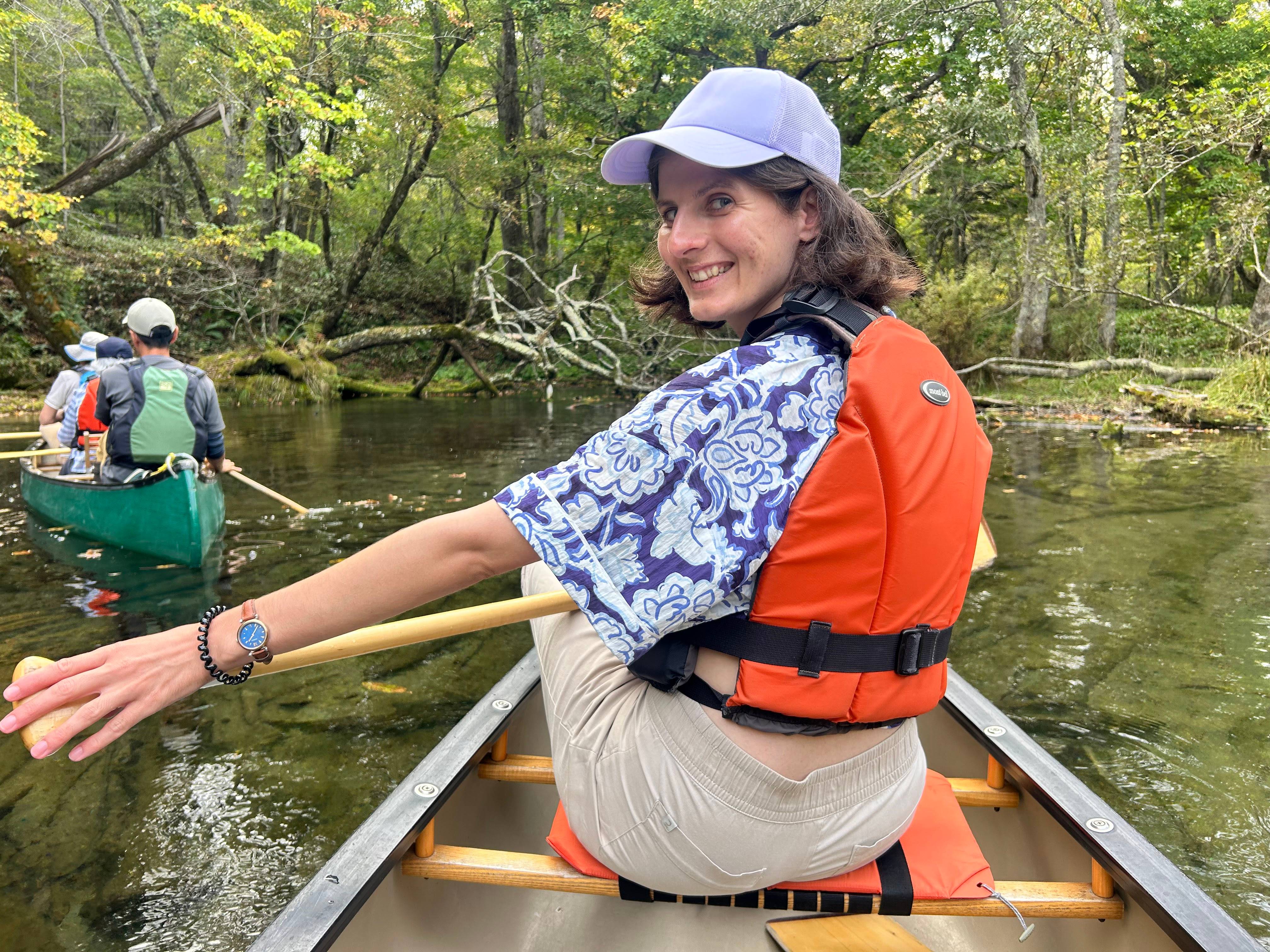A woman at the front of a canoe turns back to the camera behind her with a smile. The canoe is on a shallow river, green trees visible on the opposite bank.