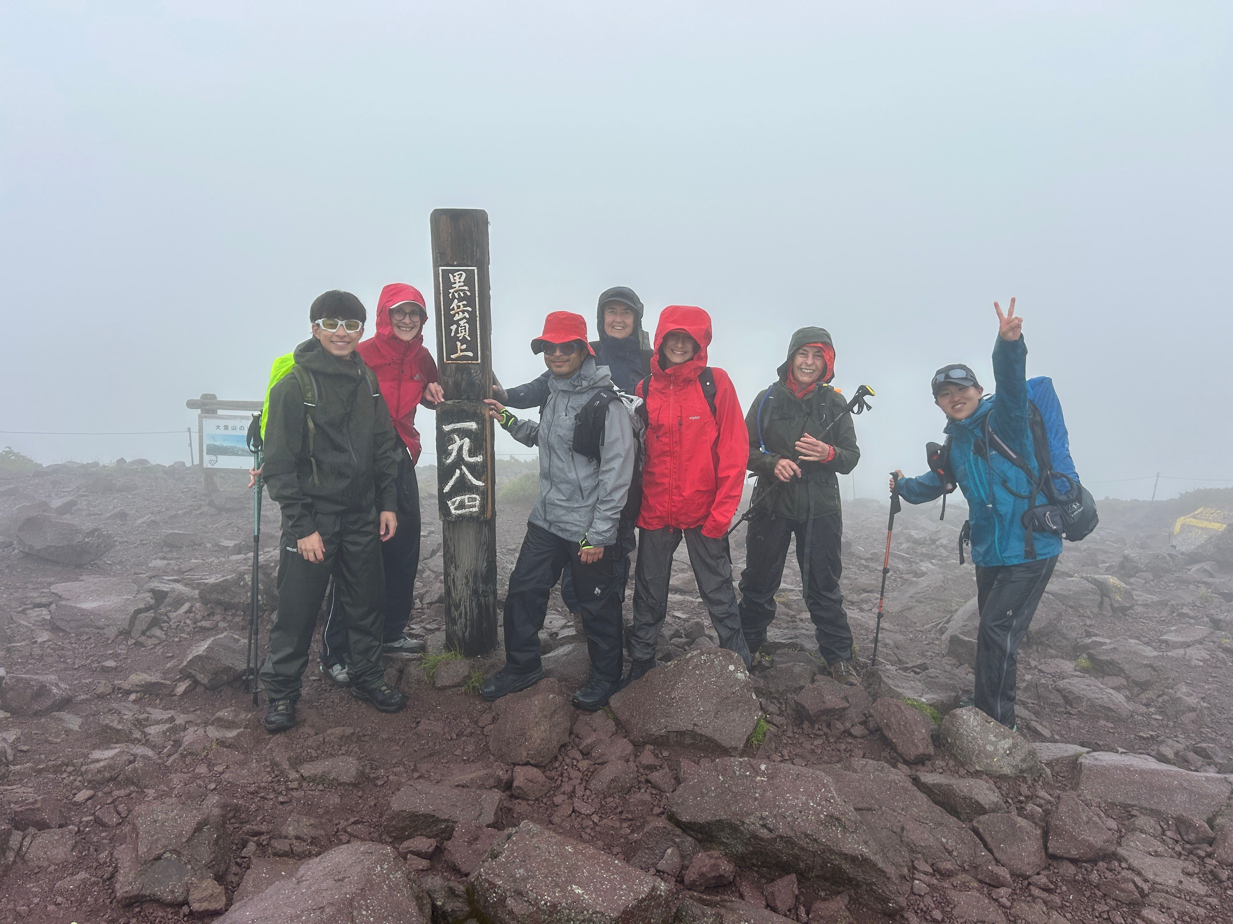 Guests smile at the camera at the summit of Mt. Kurodake in Hokkaido. A wooden pole in the ground reads "Mt. Kurodake Summit - 1,984m" in Japanese.