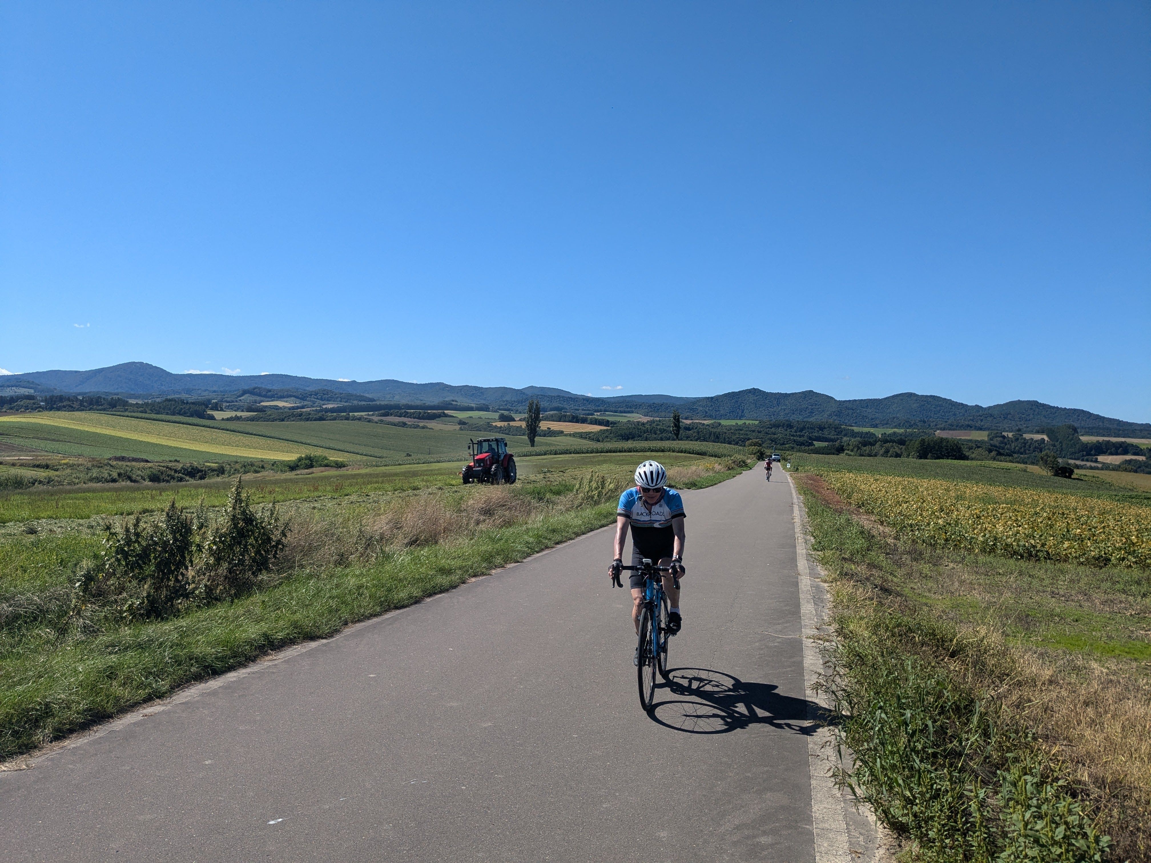A cyclist rides on a road through Biei, Hokkaido. It is a sunny day. In the background, a tractor is working in a field.