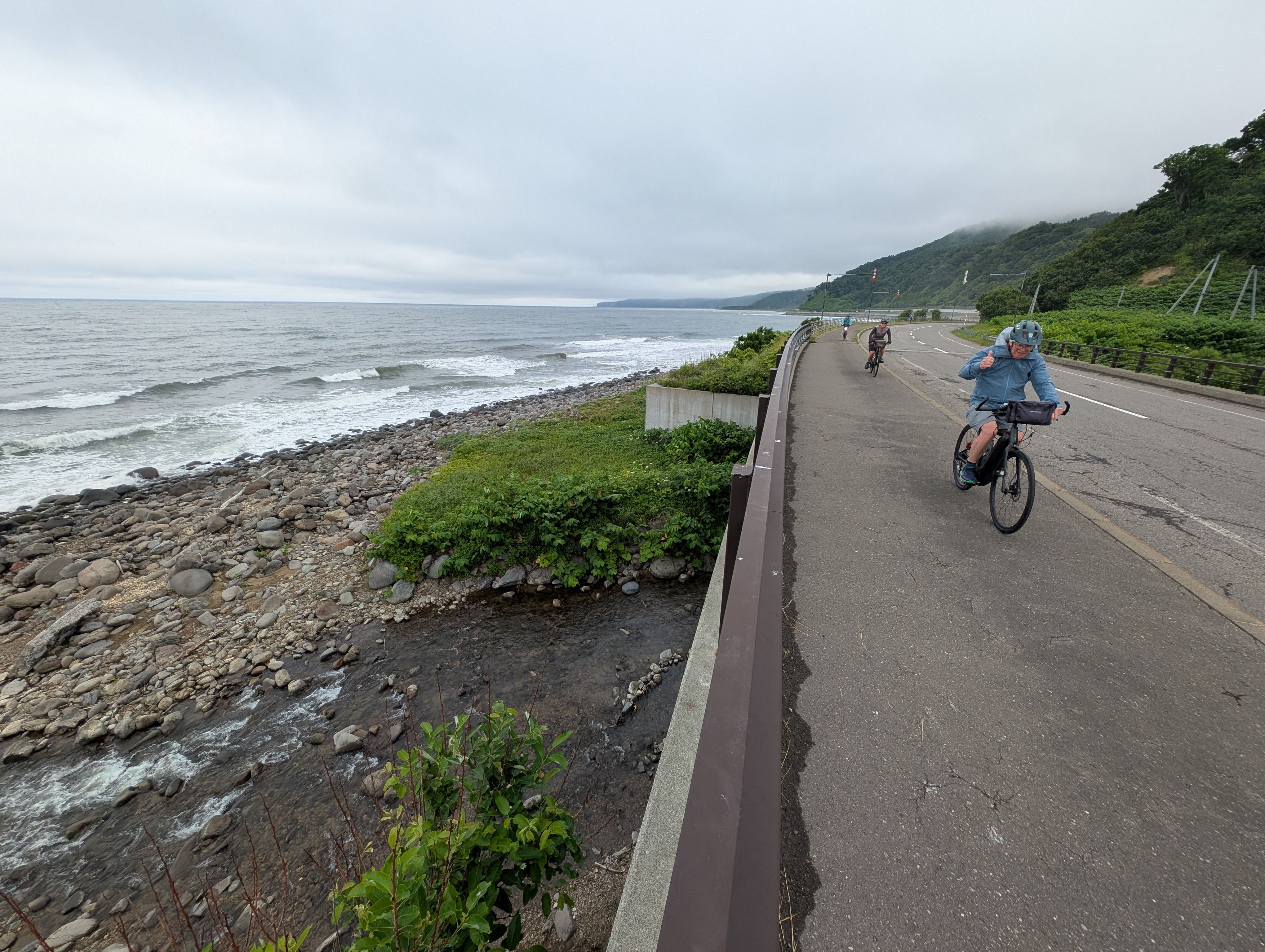 Three cyclists follow a road along the Sea of Okhotsk in east Hokkaido. It is a cloudy day and the wind appears to be high.