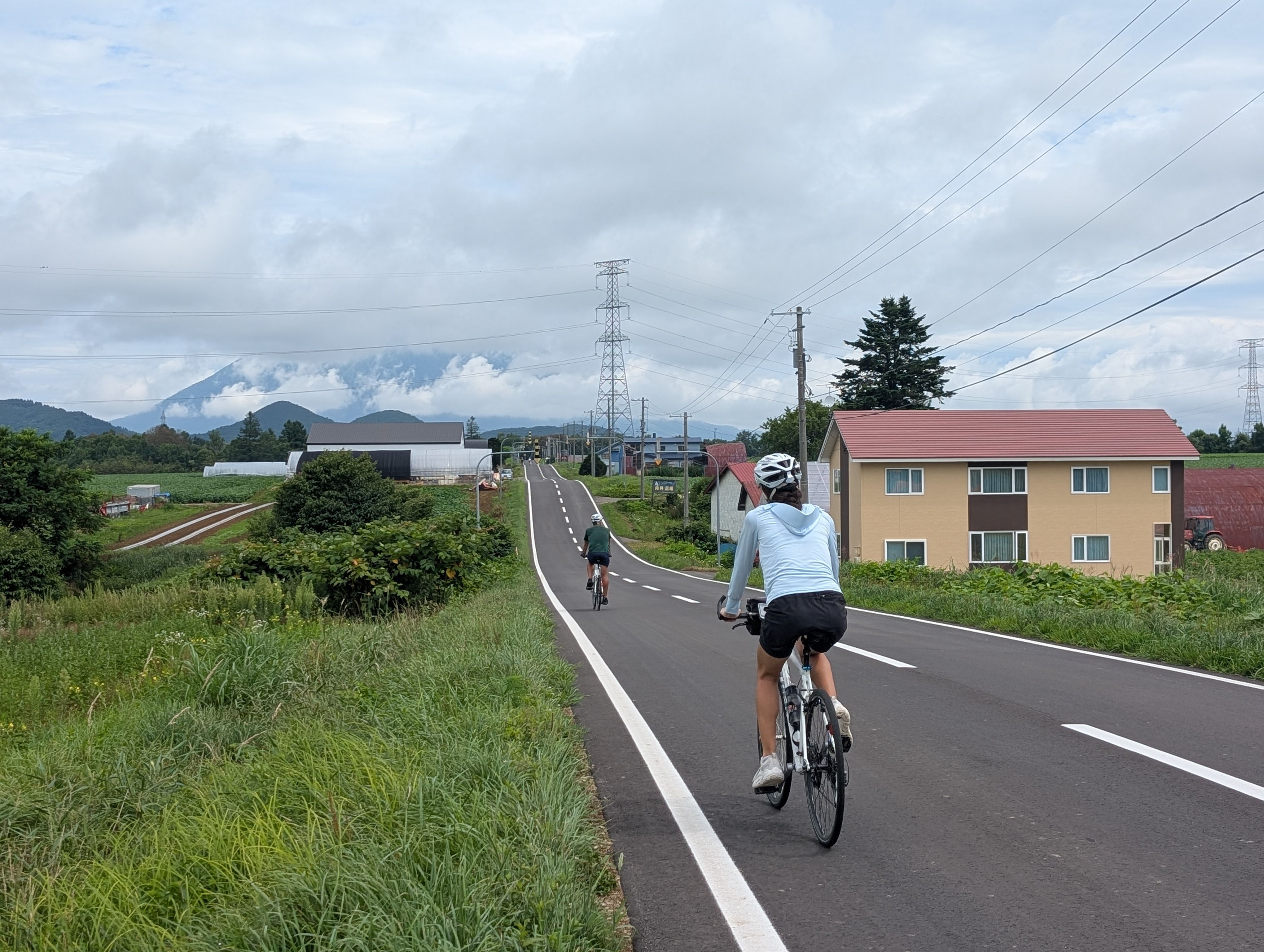 Two cyclists cycle on a gentle downhill. They are surrounded by farmland on all sides. In the distance, a mountain is starting to become visible from between the clouds.