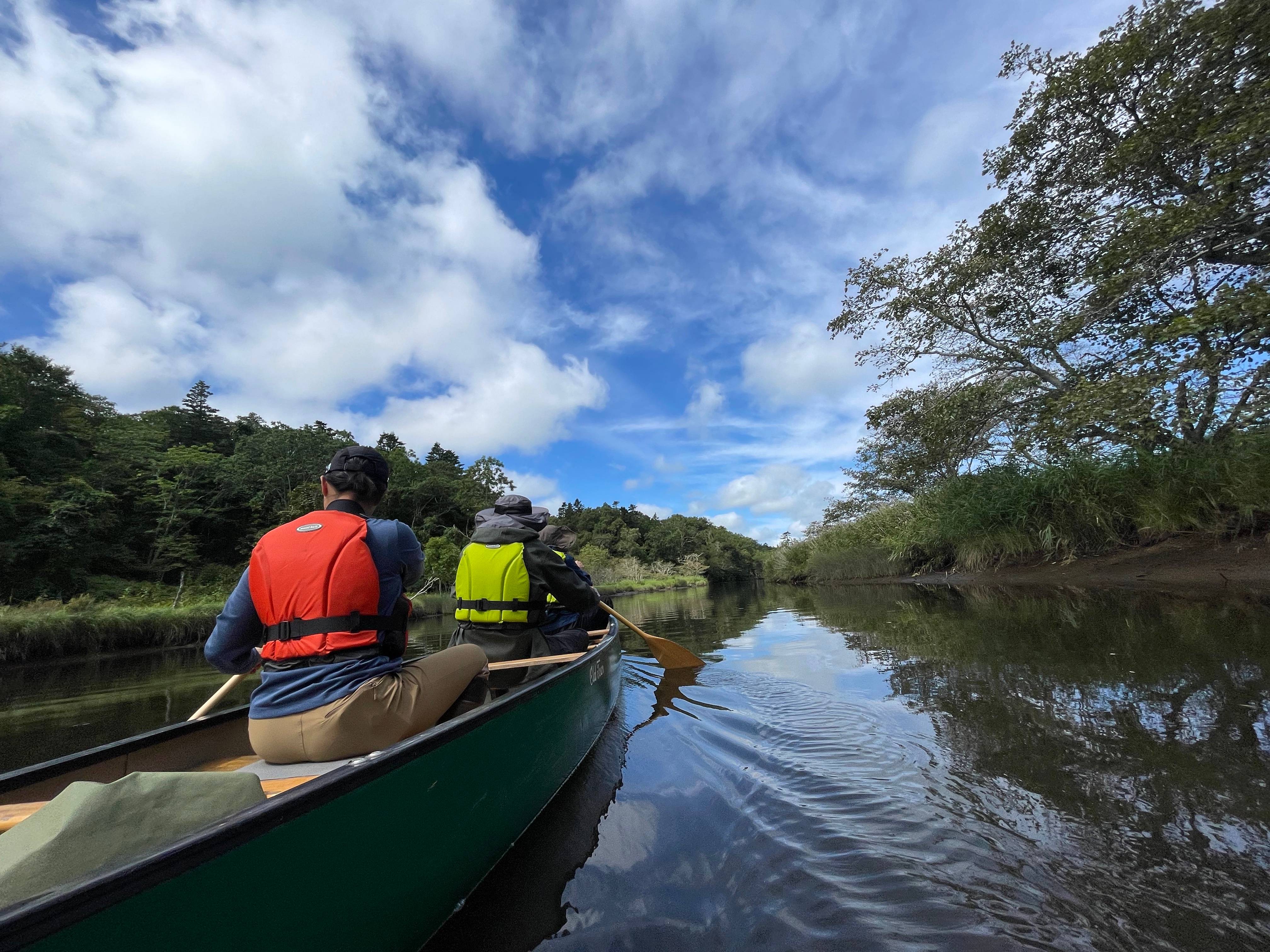 A wide-angle view taken from the back of a canoe. Two people are paddling in front and the river lies ahead. It is a beautiful day.