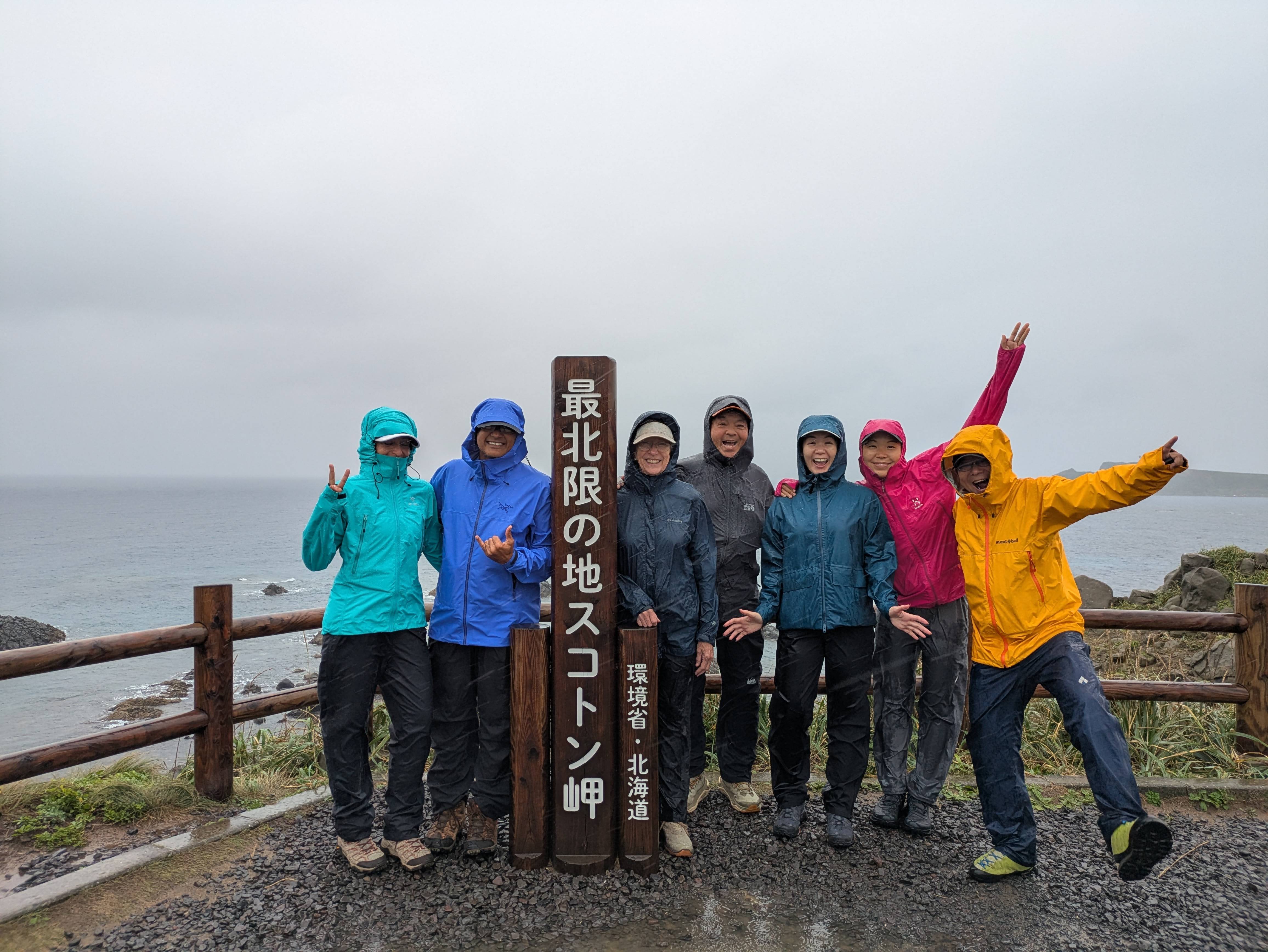 A group of hikers wear rain gear on a very damp day, standing in front of a view of Sukoton Cape. They stand either side of a sign which reads in Japanese, "Sukoton Cape, the northernmost point".