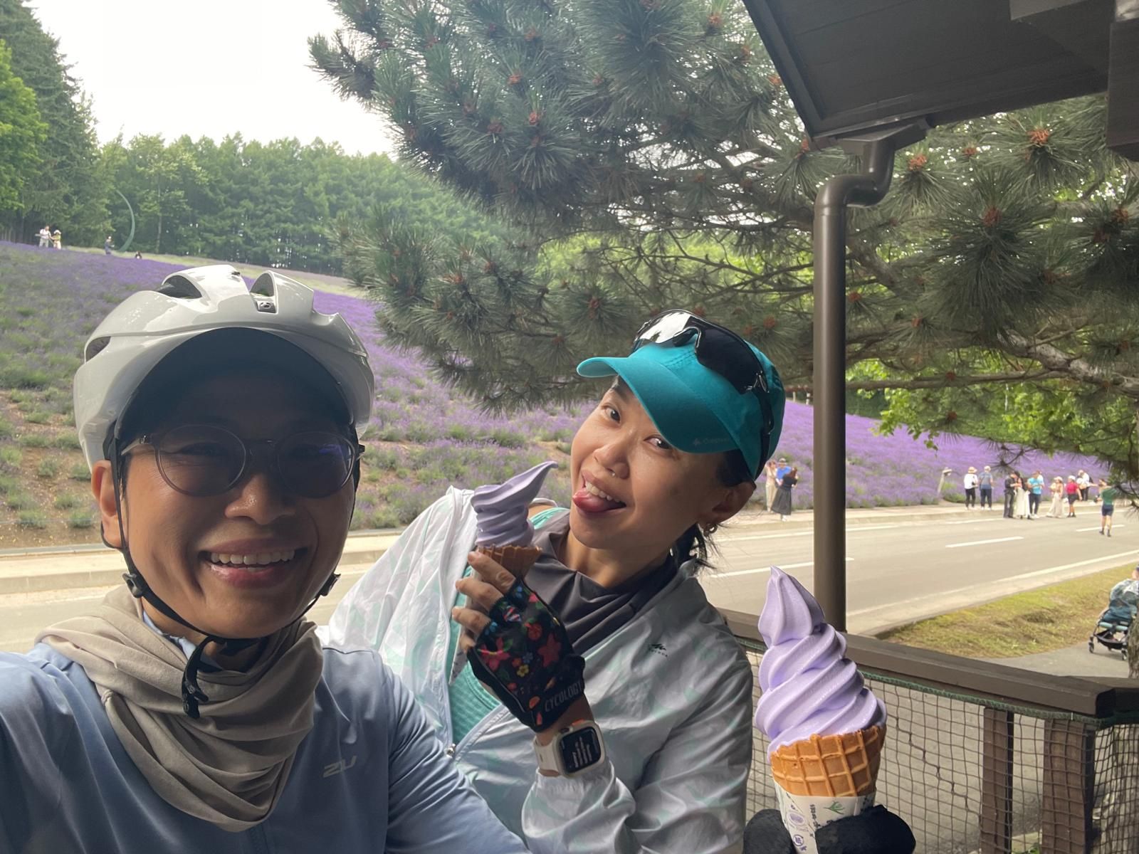 Two cyclists enjoy lavender ice cream at Farm Tomita in Furano, Hokkaido. They hold the ice cream up to the camera and smile. One cyclist is pretending to lick her ice cream.