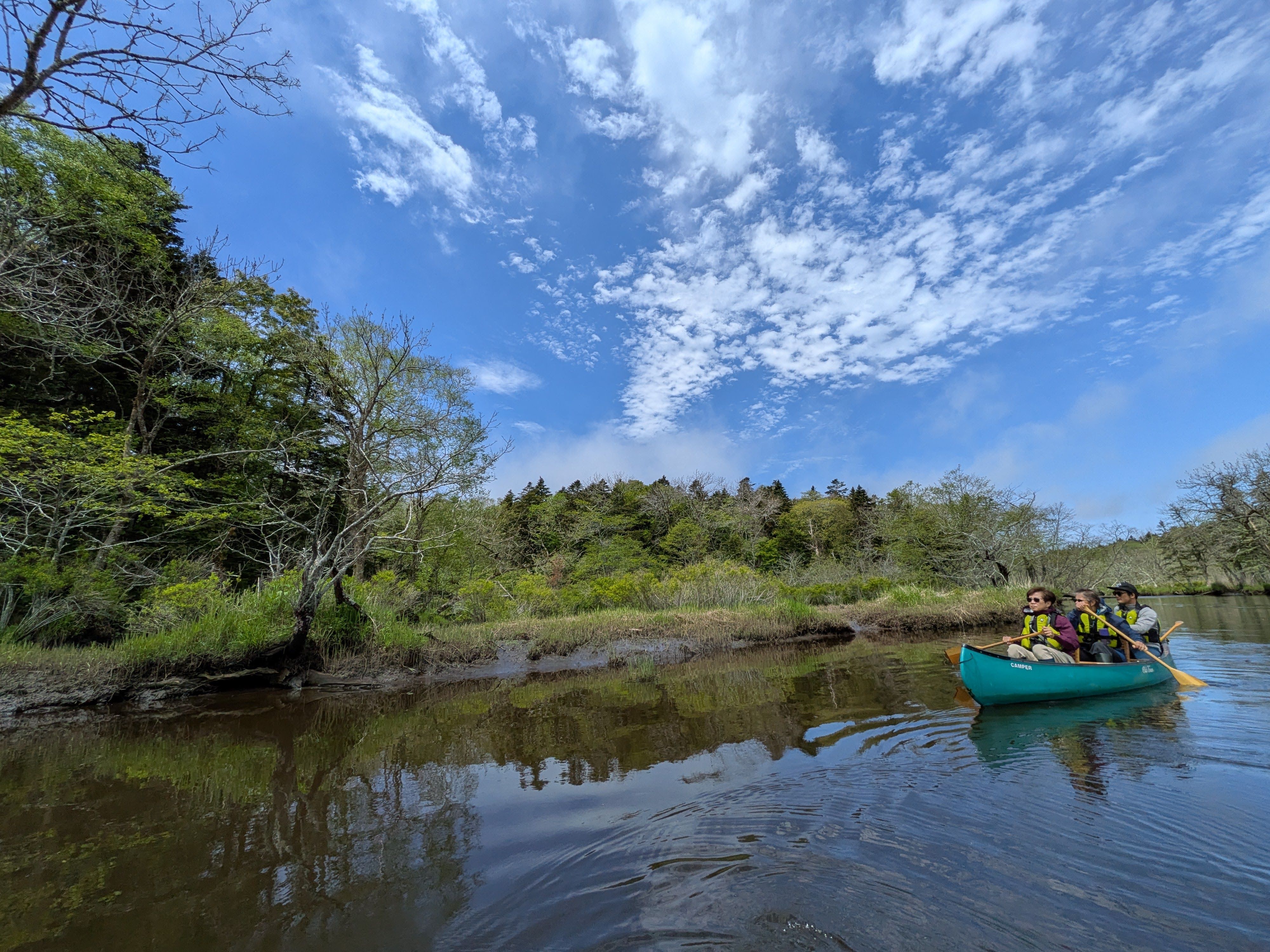 Three people paddle in a canoe on the Bettoga River, east Hokkaido. It is a sunny day and there are wispy clouds overhead. The water is quite still.
