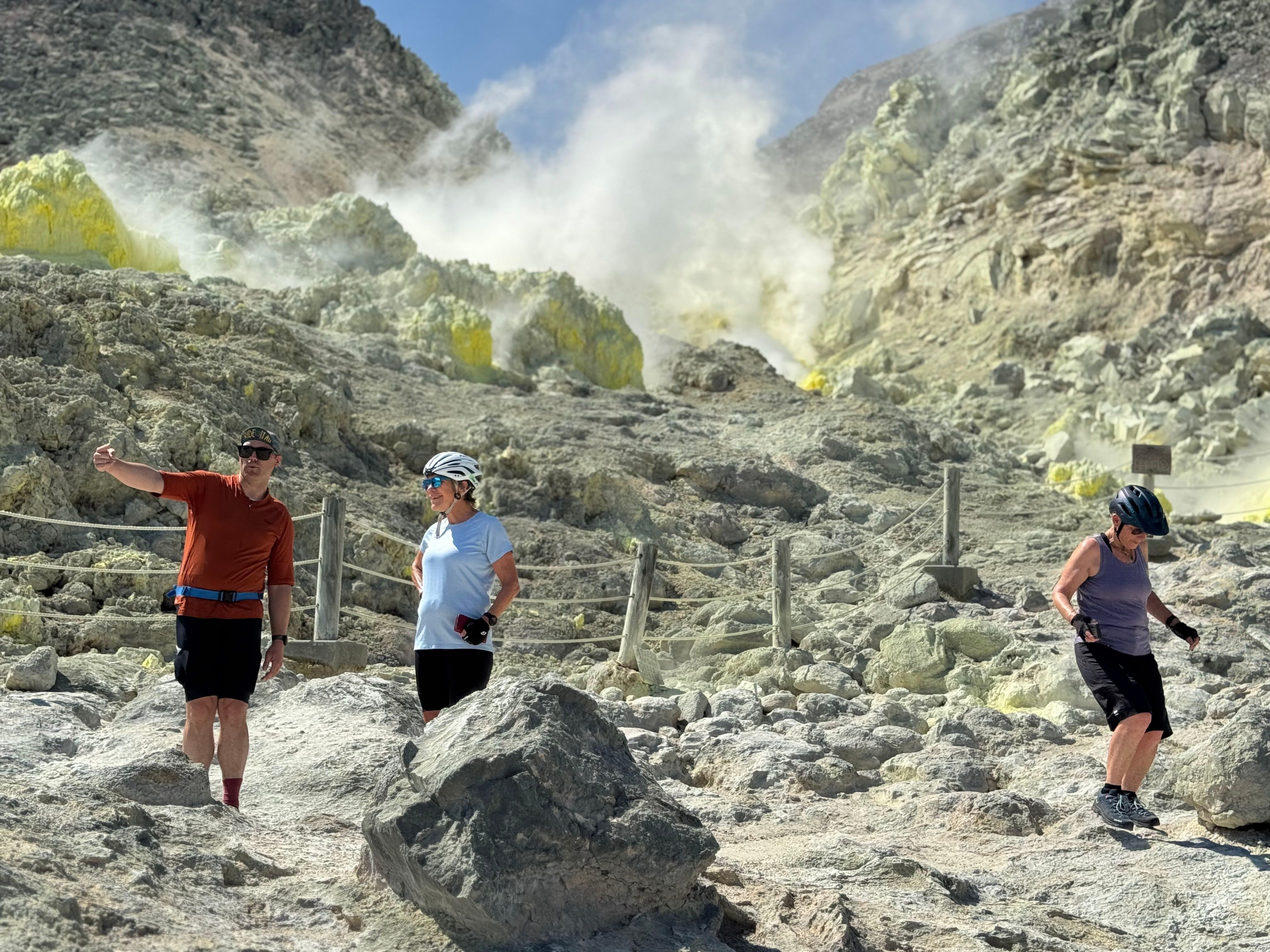 A group of cyclists gather at the base of volcanic vents on Mt. Io. Bright yellow sulphur deposits are visible in the background. One of them is taking a selfie in front of the steam.