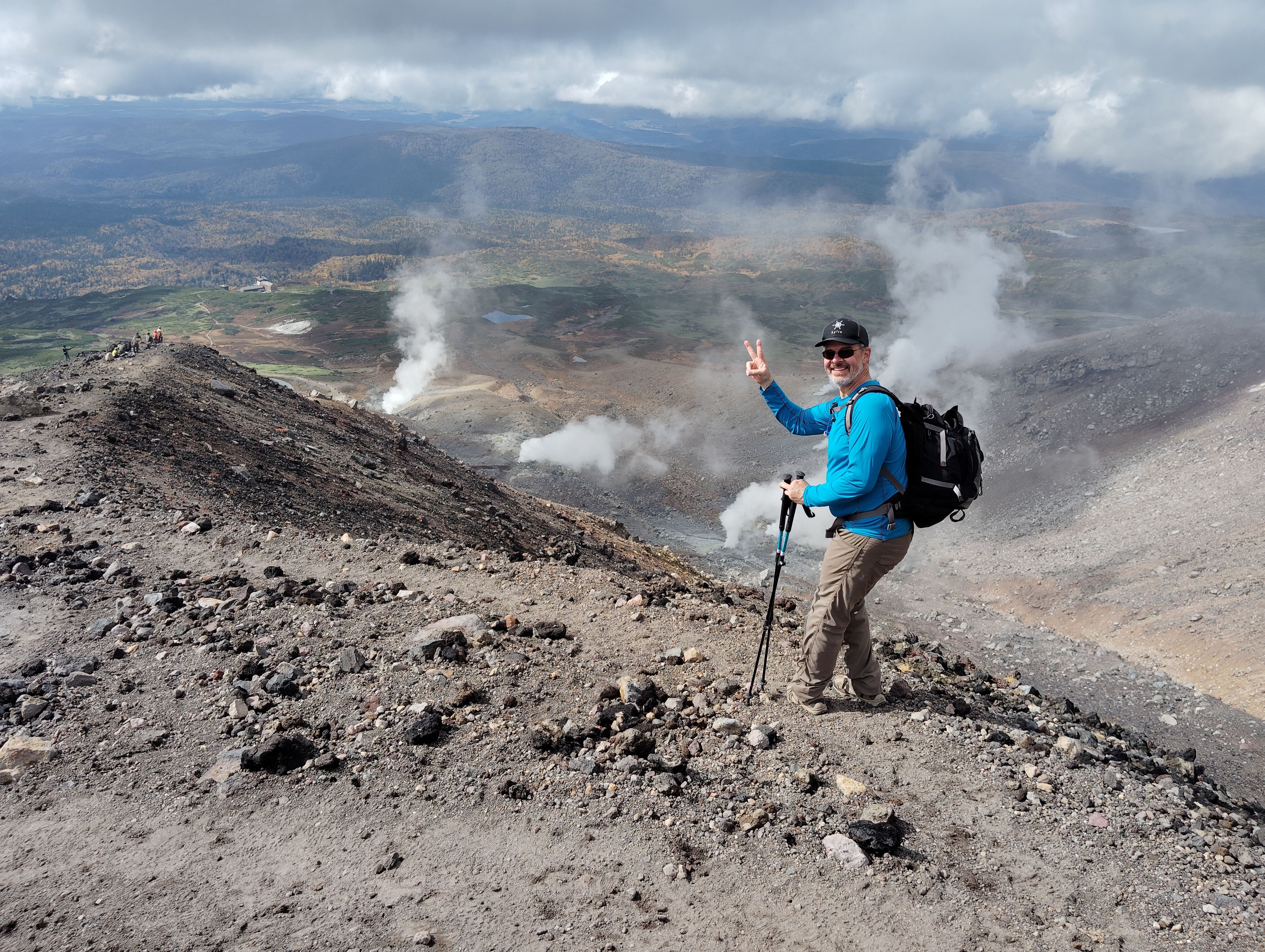 A man gives a "peace" or "victory" sign (forefinger and middle finger raised in a "V" shape) to the camera on a rocky mountain trail. Behind him, volcanic fumaroles are visible in the distance, sending up columns of steam.