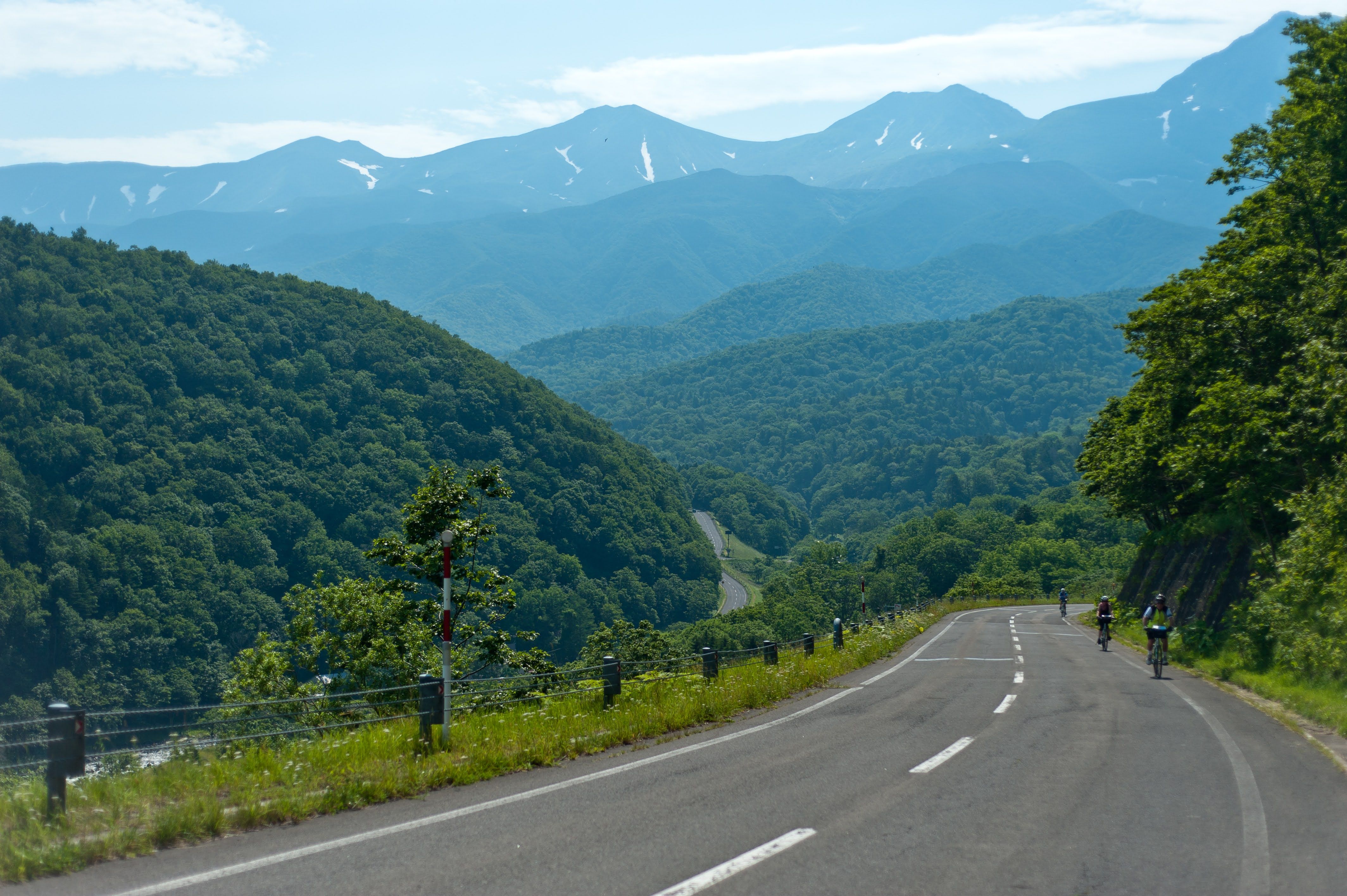 A scenic view of people cycling on a winding road through the green mountains of Shiretoko, with snow-patched peaks in the background.