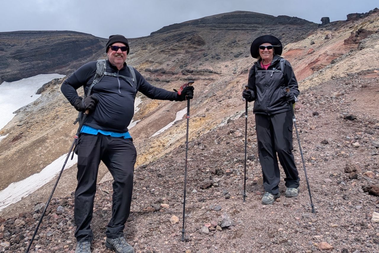 A smiling man and woman stand on a rocky, volcanic trail while hiking Mt. Asahidake in Hokkaido. Both wear black hiking gear, hats, and sunglasses, holding trekking poles. The rugged mountain backdrop features steep, colourful slopes and patches of snow.