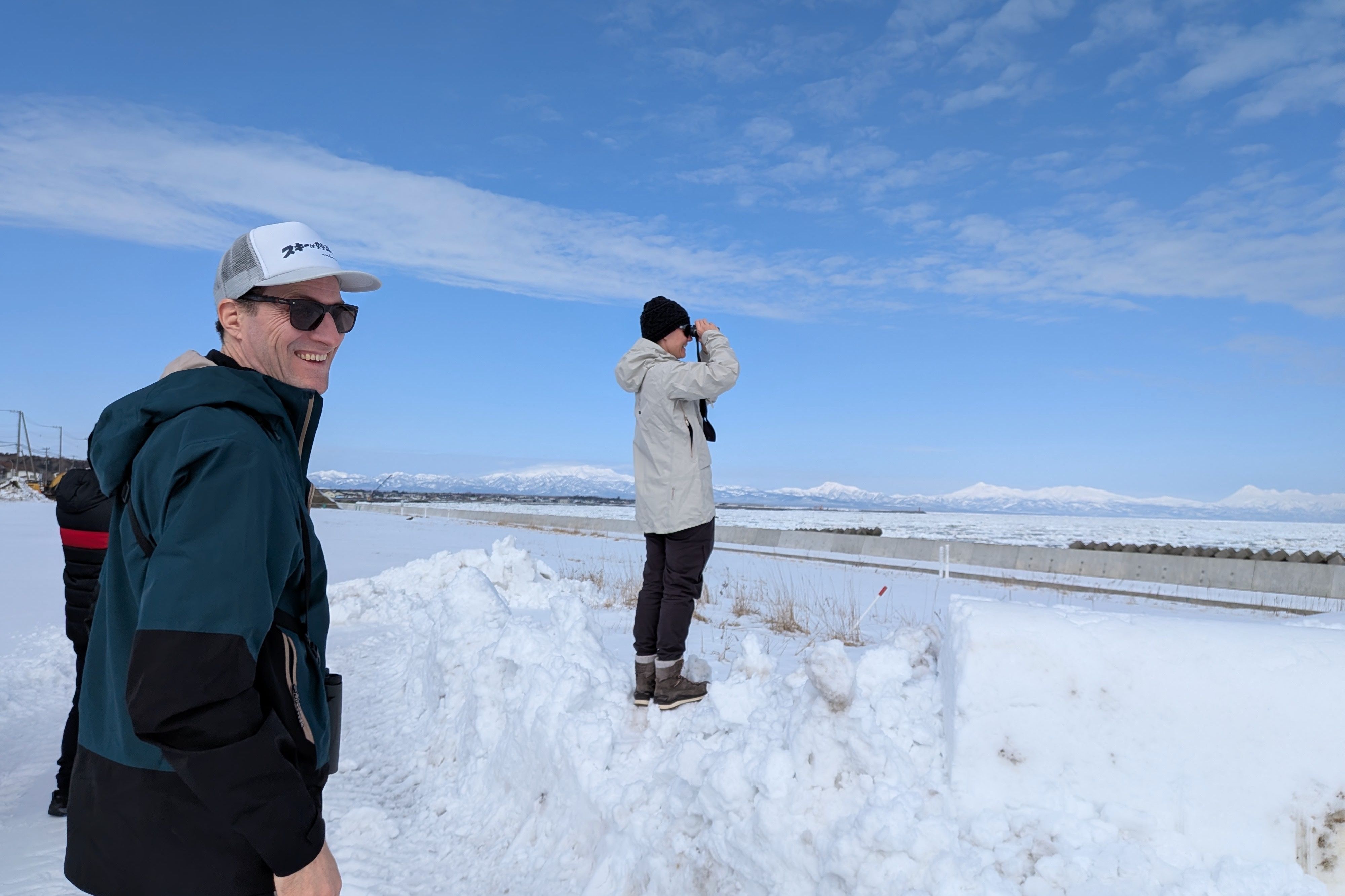 A smiling guest in winter gear looks happily at the camera while another guest stands on a snowbank behind him, using binoculars to admire the snowy landscape and snow-capped mountains across the frozen Nemuro Strait near Shibetsu, Hokkaido.