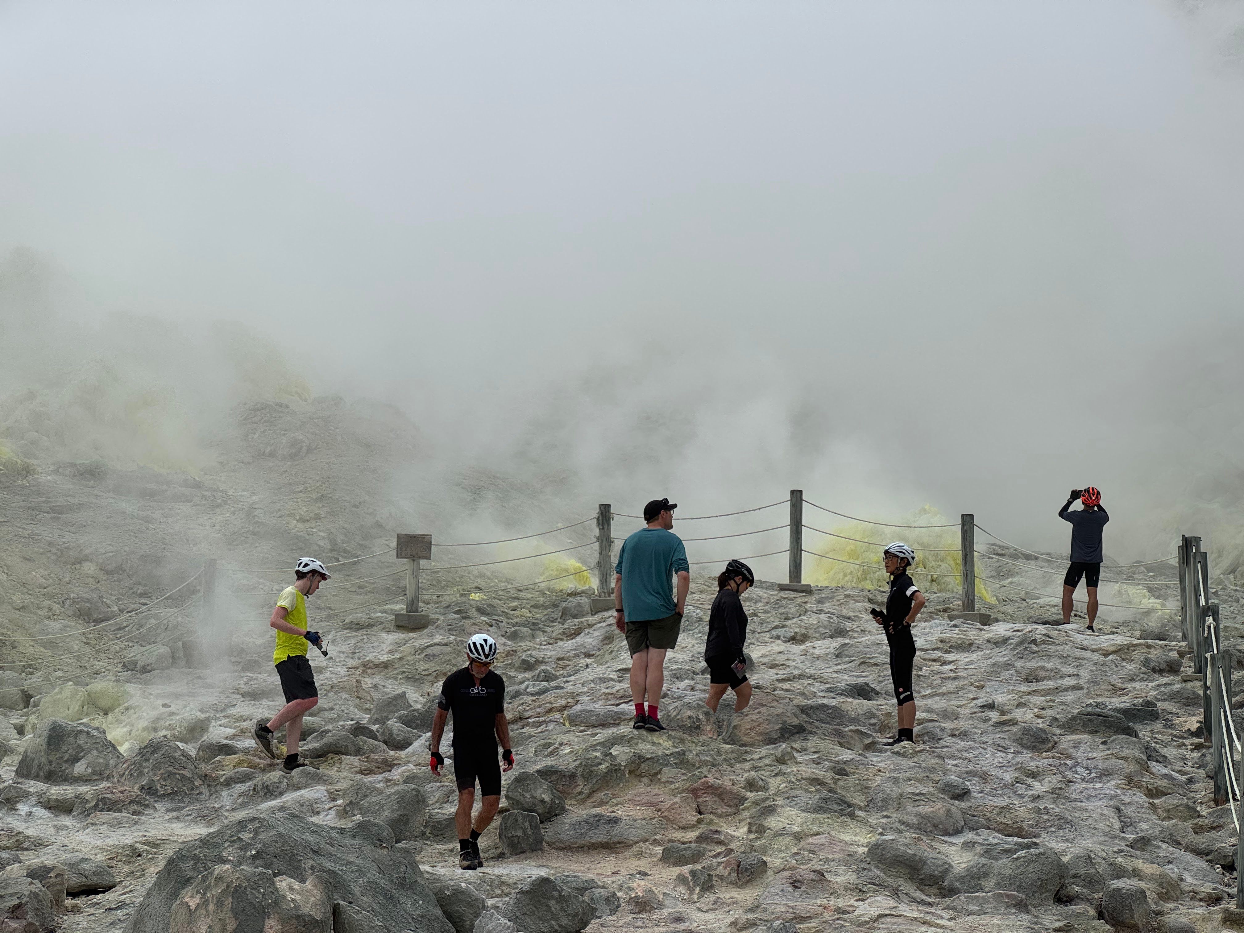 A group of cyclists gather around the steaming crater of Mt. Io, an active volcano in Hokkaido. There is steam rising all around them and rocks that have become completely yellow with sulphur deposits.