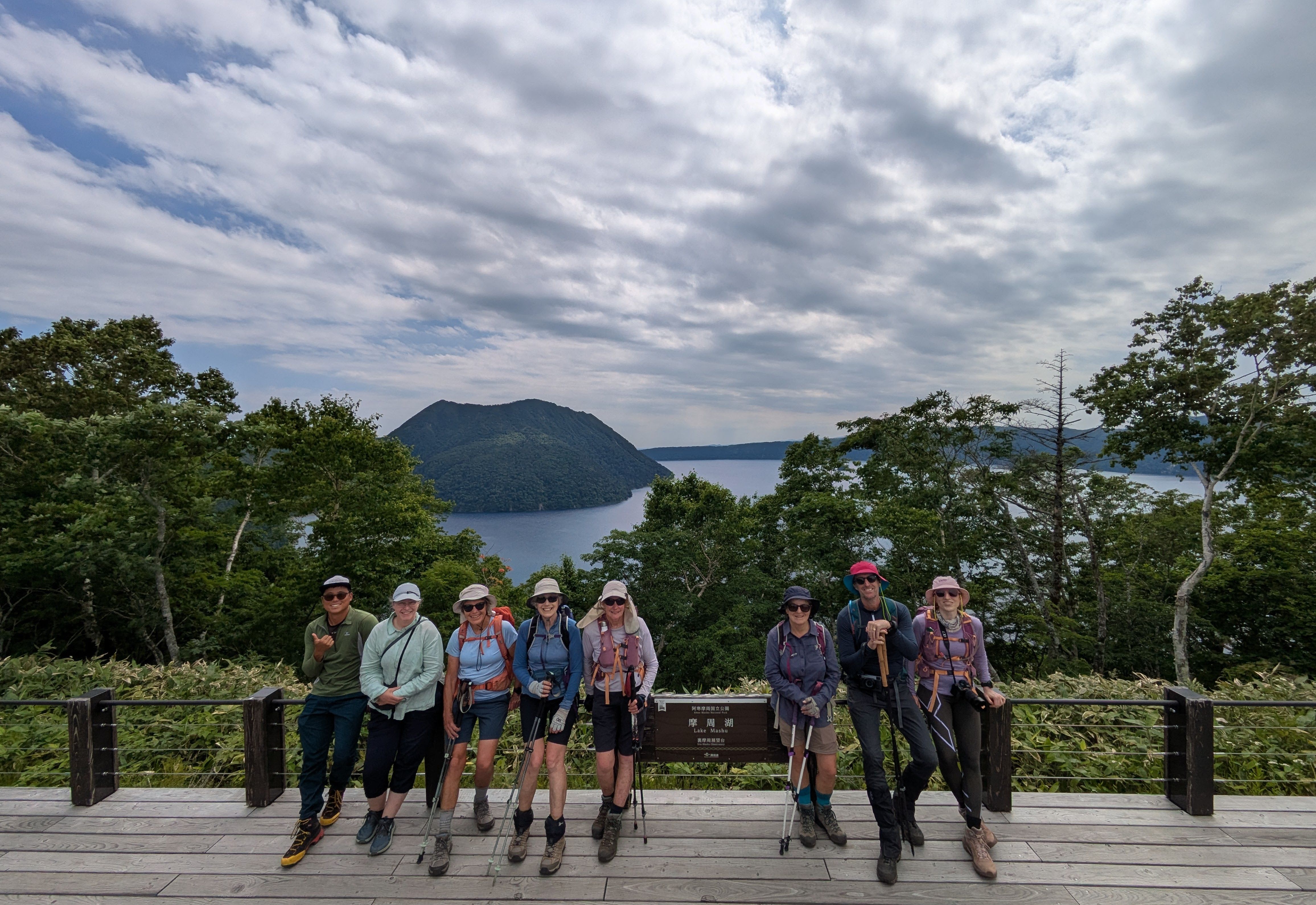 A group of hikers stand around a sign at a lookout over Lake Mashu. The lake is visible behind the group.