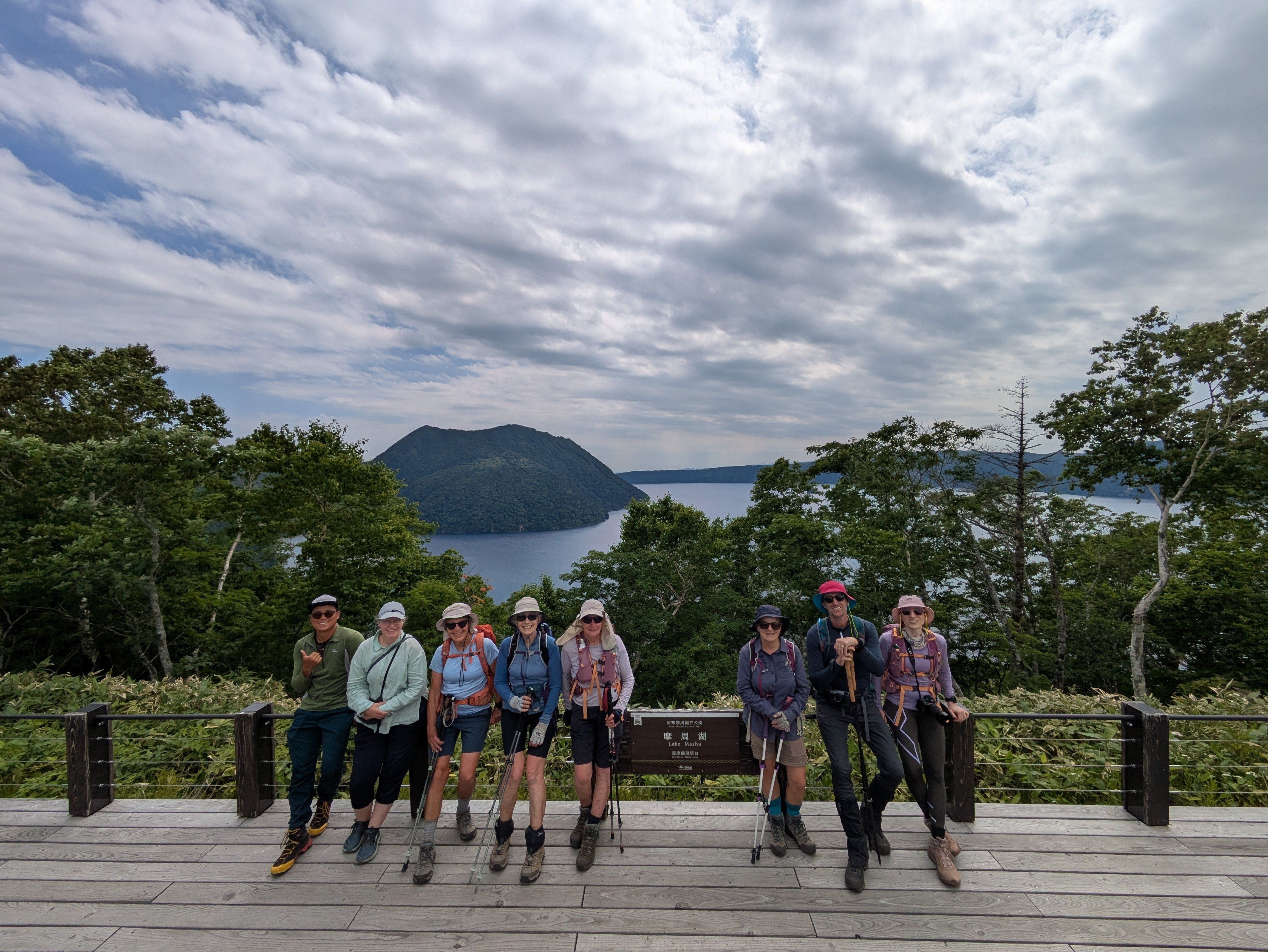 A group of hikers stand around a sign at a lookout over Lake Mashu. The lake is visible behind the group.