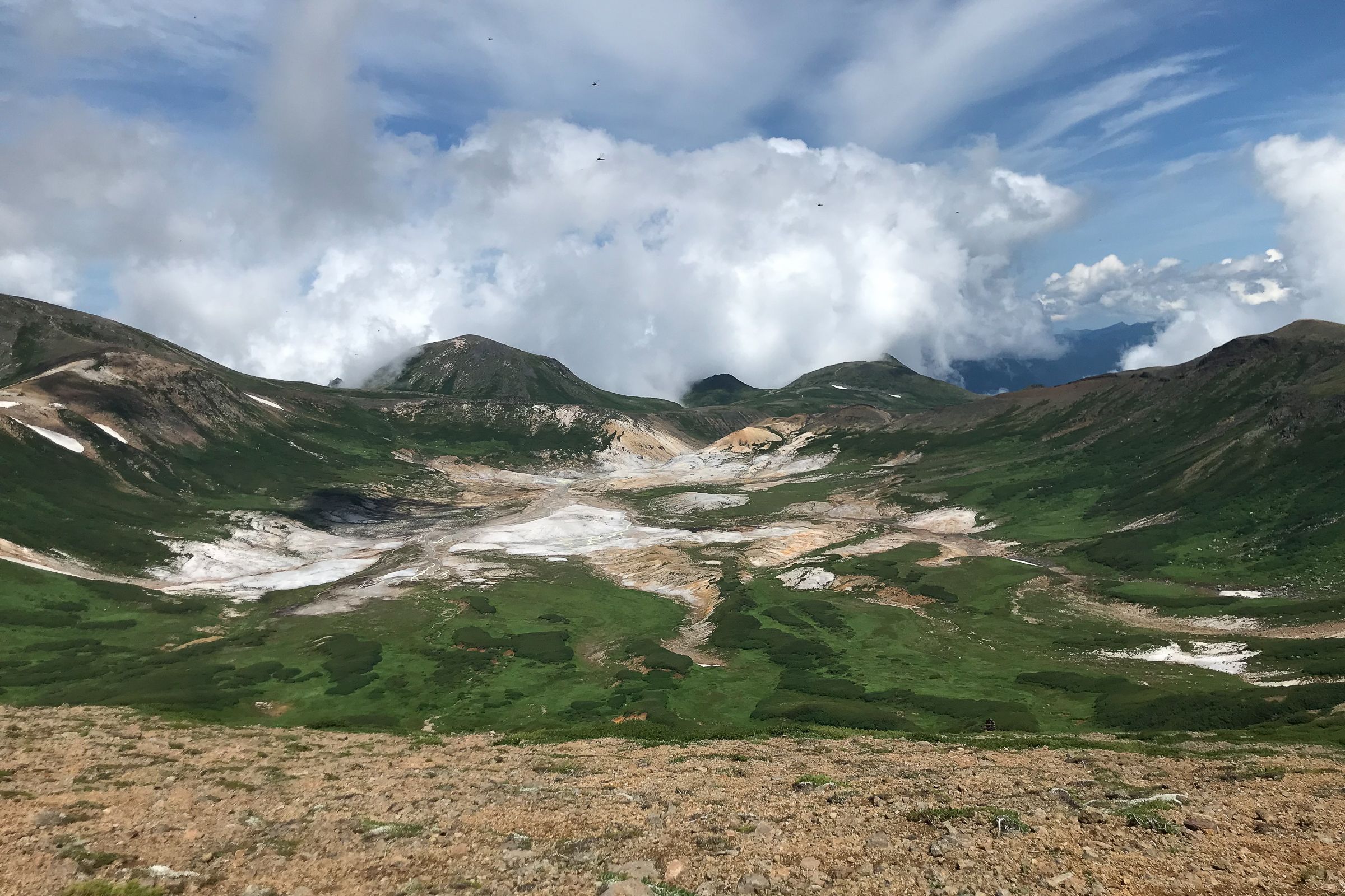 The vast expanse of the Ohachidaira caldera stretches away from the camera. In the background white clouds brew in a blue sky surrounding the high peaks of the Daisetsuzan