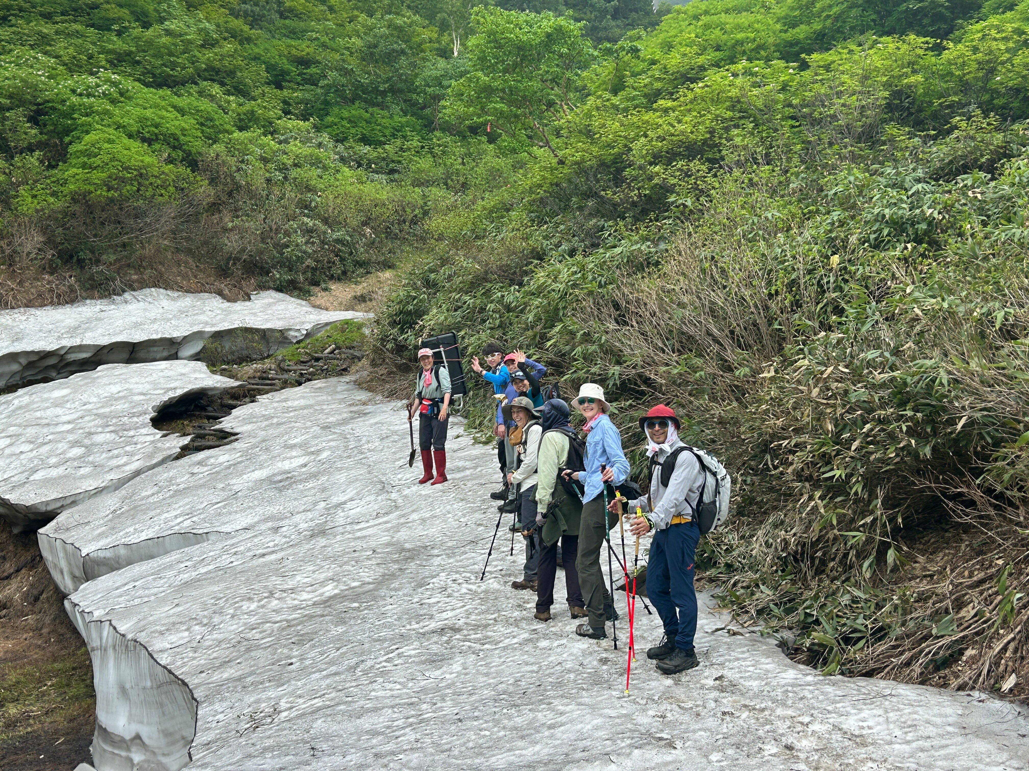 A group of hikers stand on a snow field, looking back at the camera and waving.