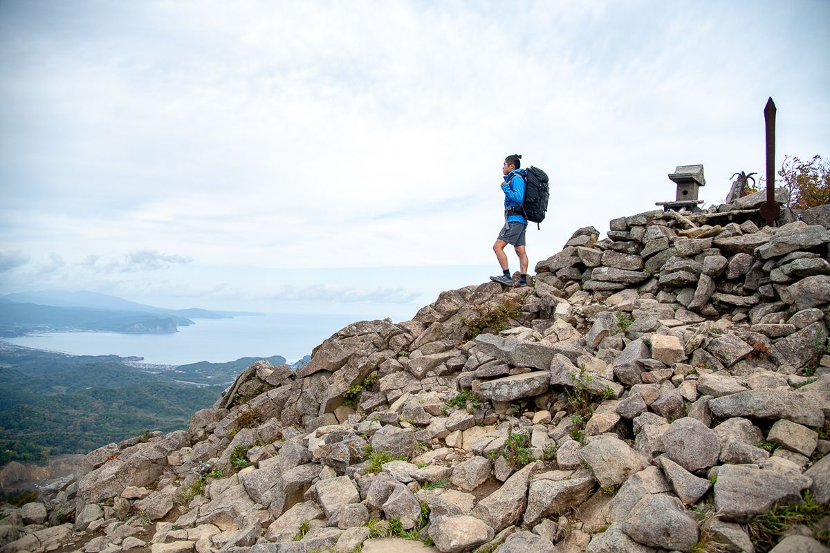 A hiker with a large pack stands on the rocky summit of Mt. Shioya-Maruyama, next to a small stone shrine. He looks out over a sweeping coastal view of a bay and the sea near Otaru.