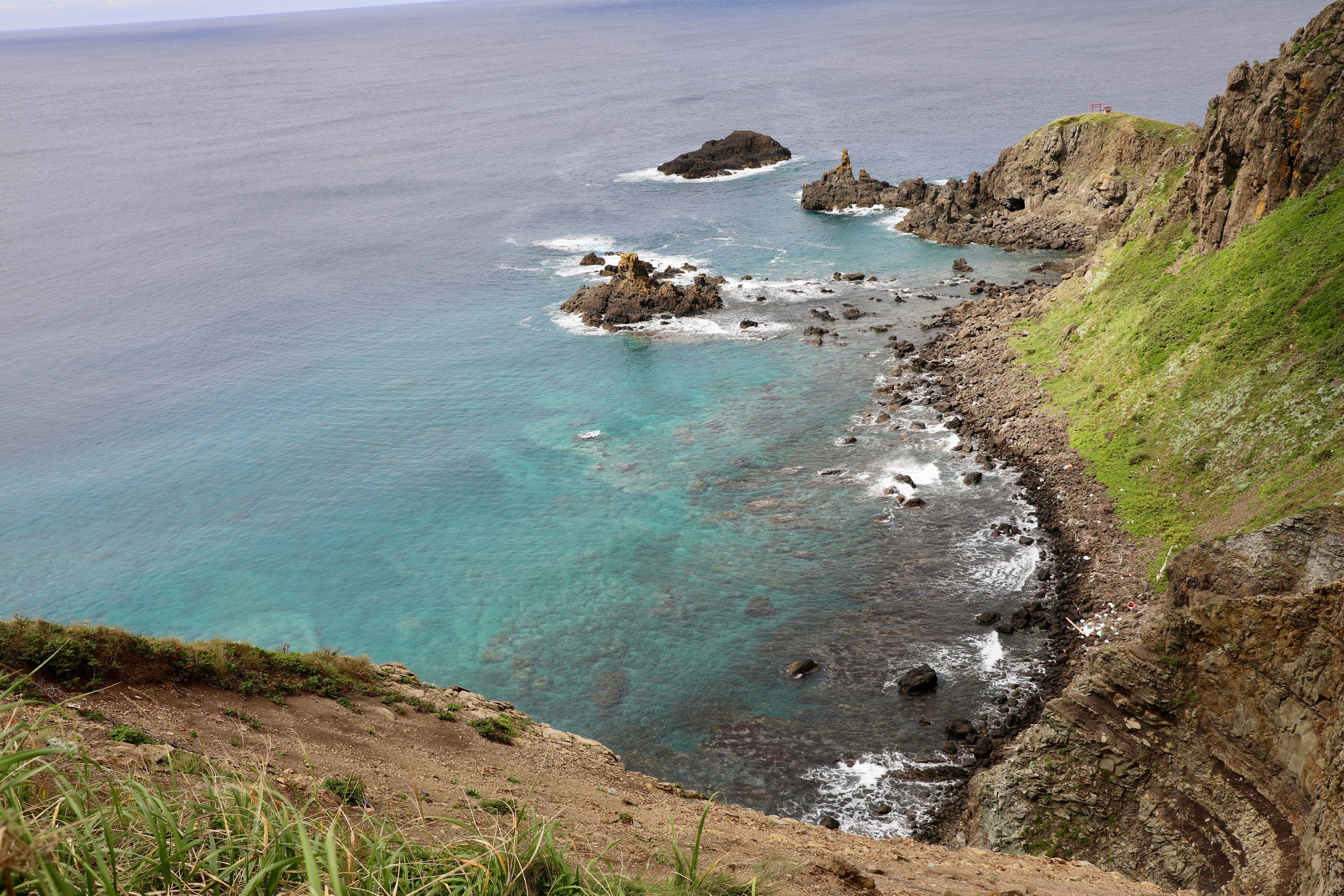 Turquoise water laps up against the rugged and rocky shoreline of Rebun Island in Hokkaido. There is a distinct vertical rock sticking up and the end of the headland.