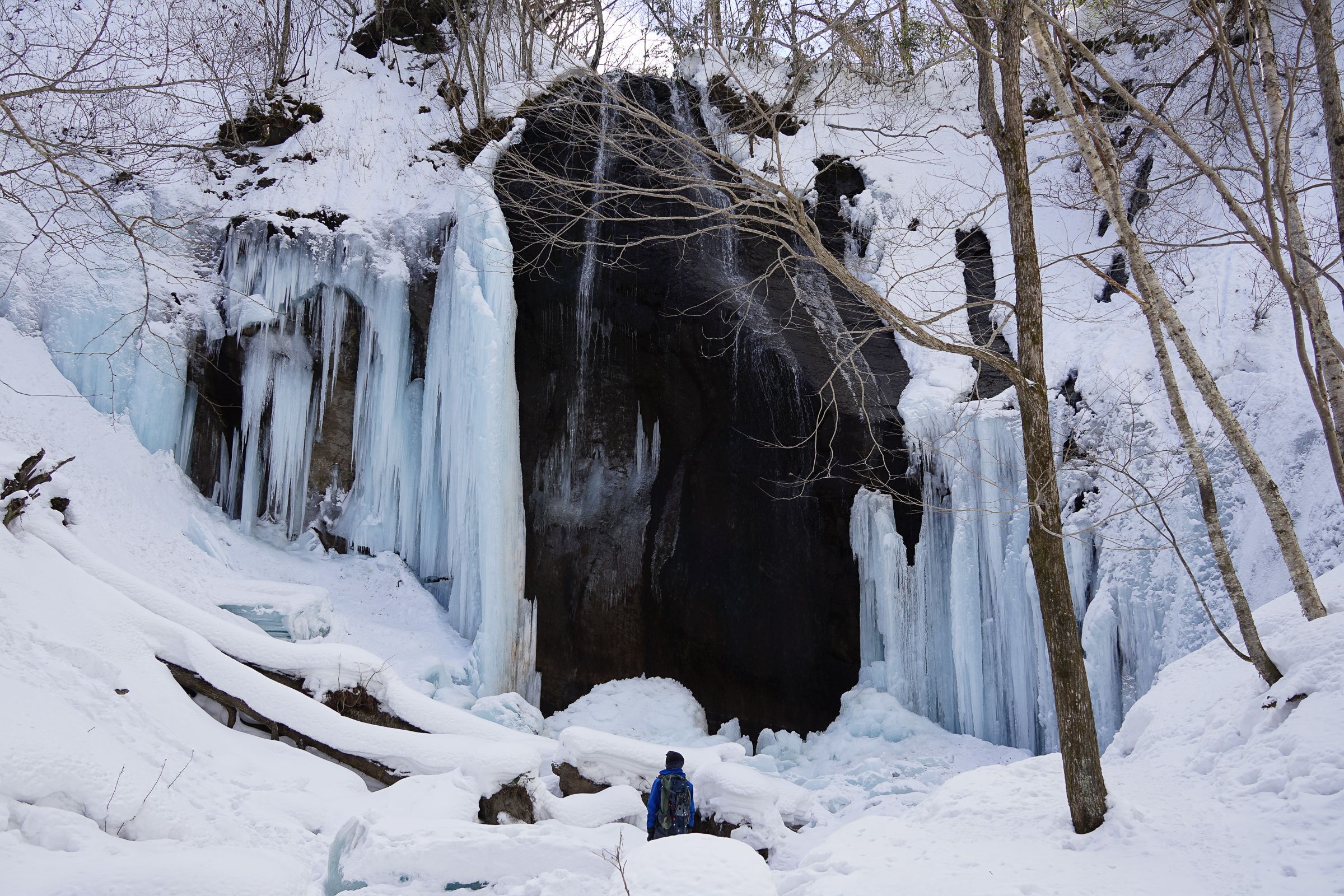 A hiker in a blue jacket and snowshoes stands before a massive frozen waterfall in Shiraoi, Hokkaido. The winter scene features towering pale ice pillars and sharp icicles framing a dark rock face, surrounded by deep drifts of white snow.