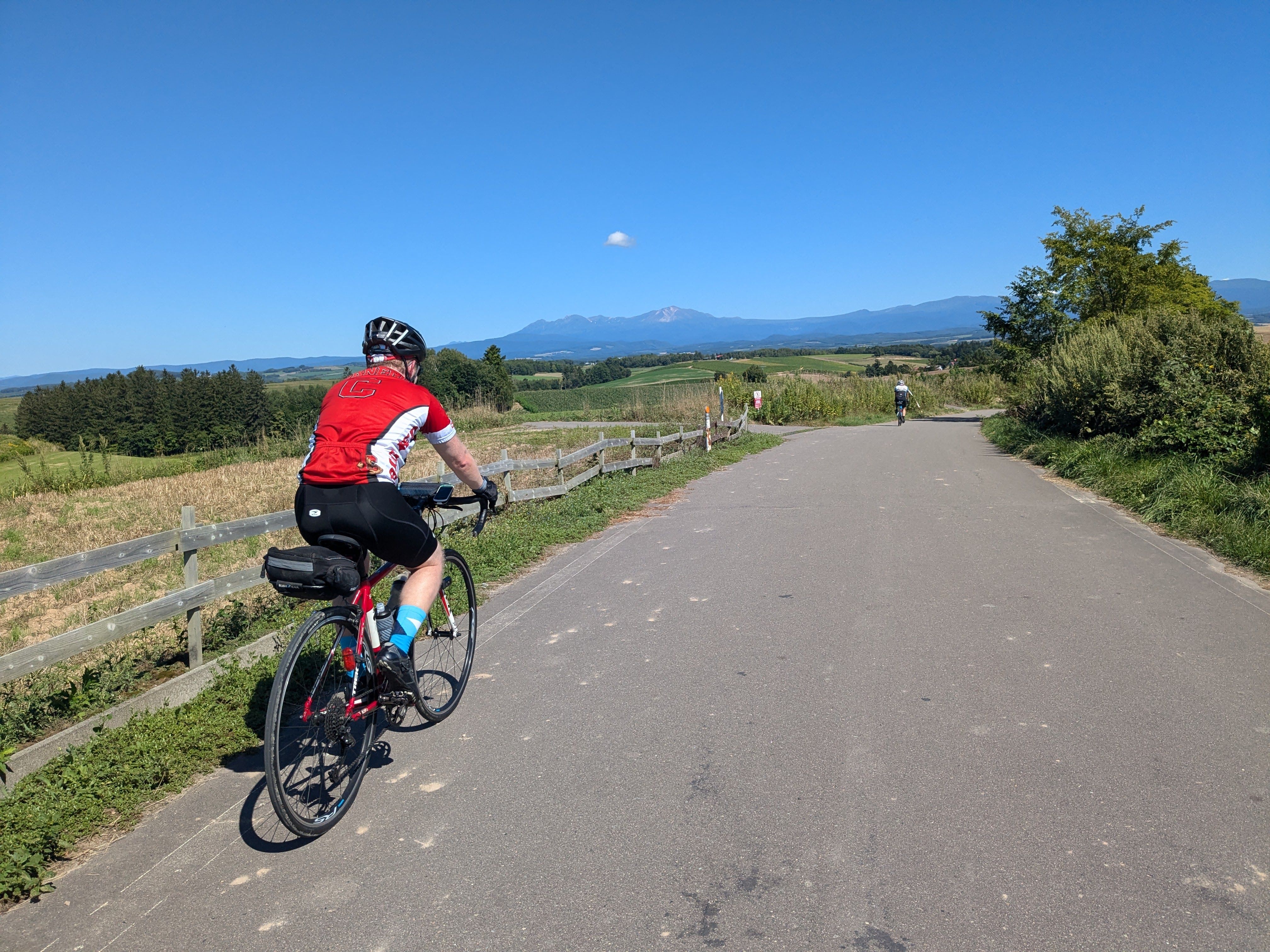 A cyclist climbs a hill alongside a fenced field in Biei, Hokkaido. Mt. Asahidake, the tallest mountain in Hokkaido, is visible in the distance.