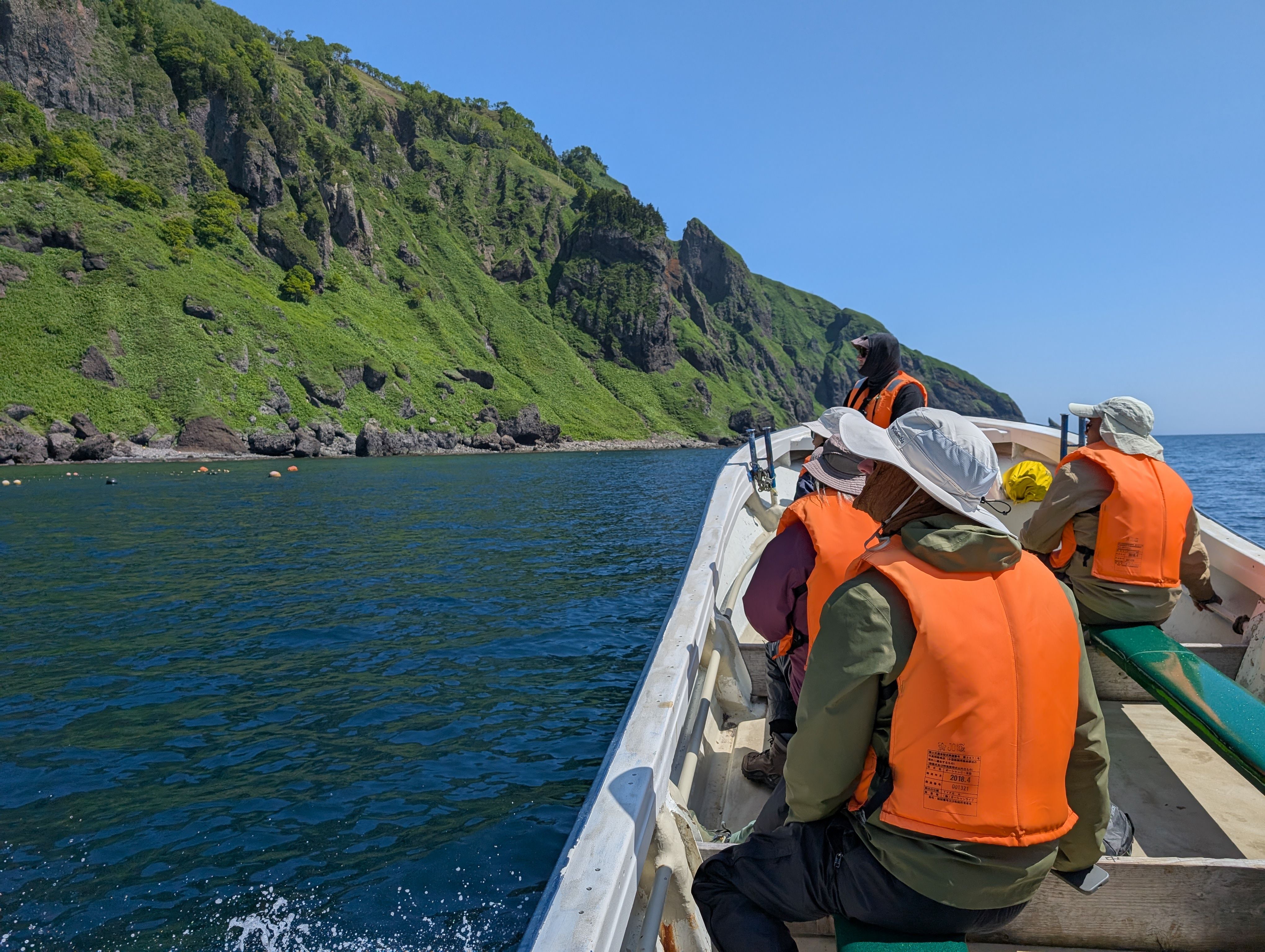 A group of people in life jackets sit in a small fishing boat along the shore of the Shiretoko Peninsula. It is a sunny day. The sea is blue and the cliffs of the peninsula are a striking, leafy green.