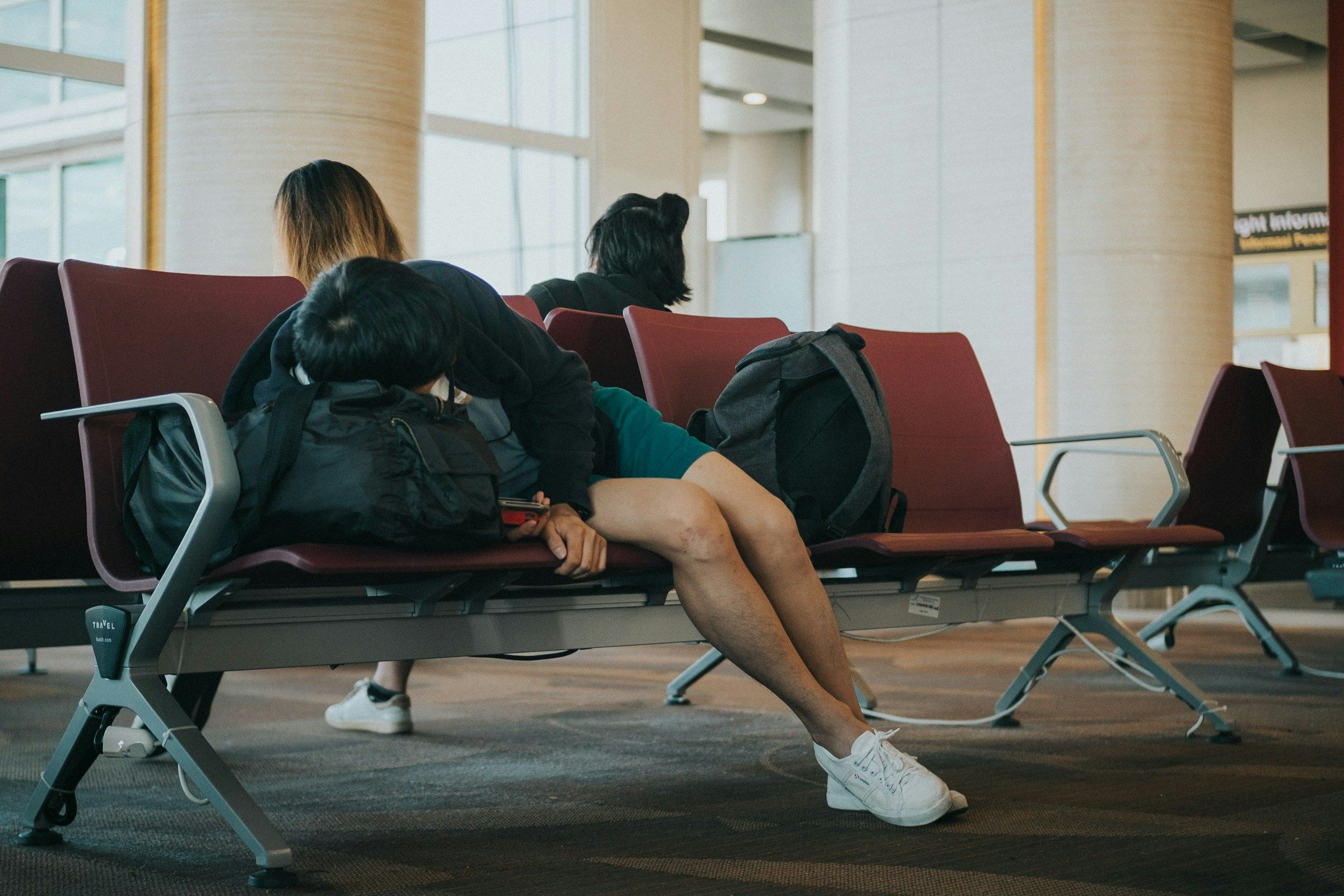 A tired traveler sleeps slumped over their backpack while waiting in a row of airport terminal chairs.