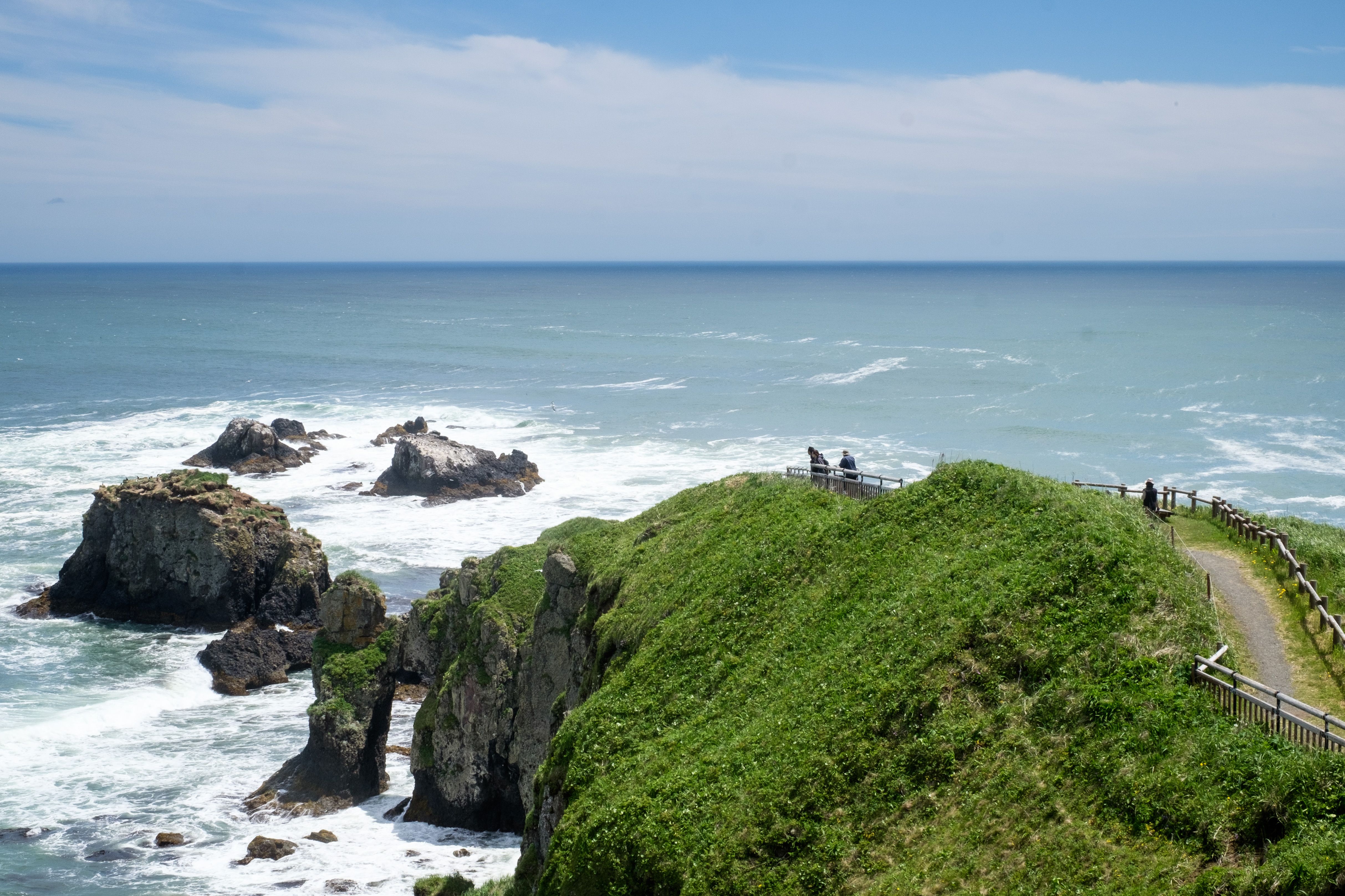 A group of walkers make their way out to a wild headland. The cliffs are surrounded by white water as waves crash into shore.